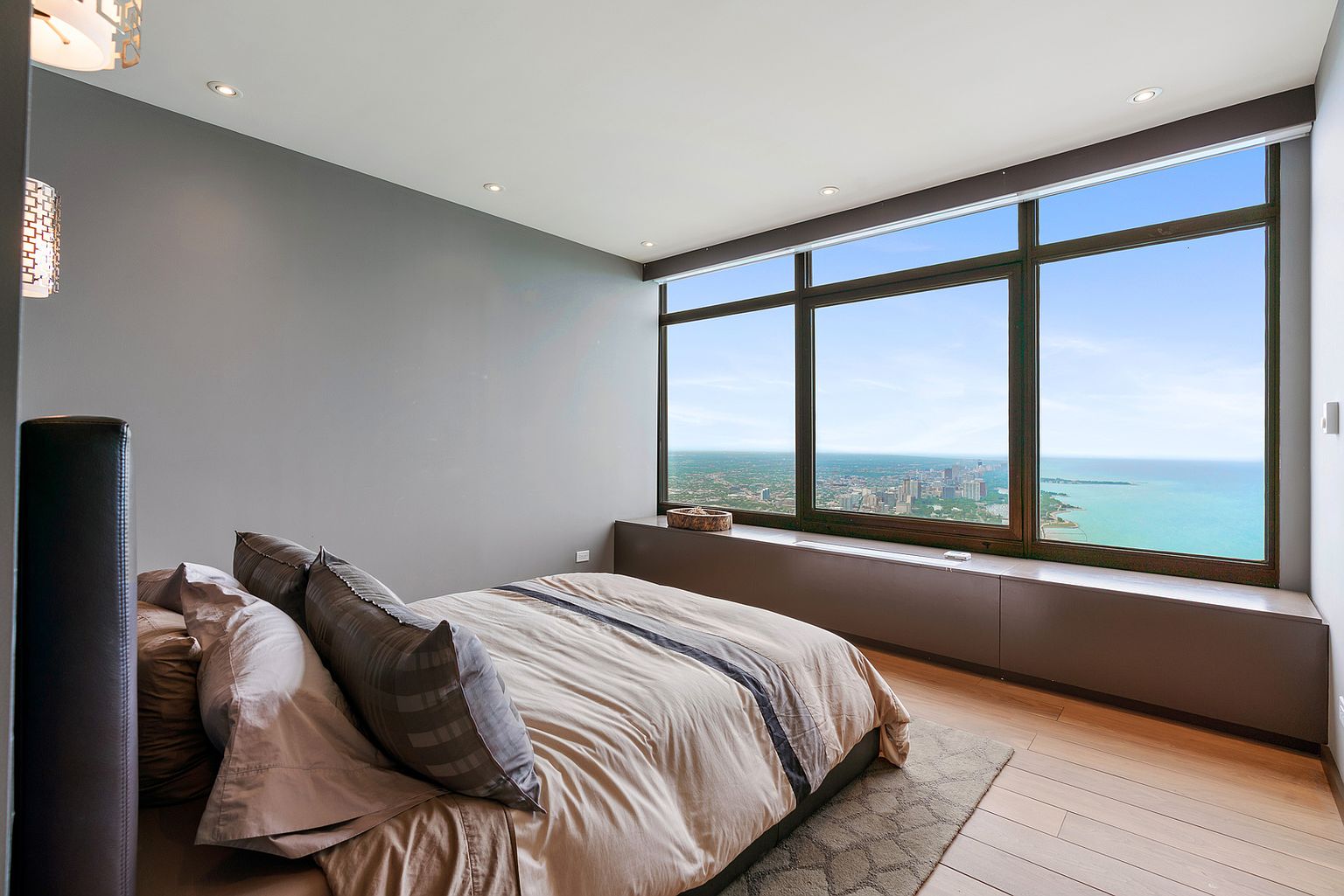 This modern primary bedroom features a neutral color palette and a low-profile bed centered in the room. A large window wall provides an expansive, panoramic view of the city skyline and the adjacent body of water. The space is finished with light wood flooring and a minimalist built-in ledge beneath the windows, creating a sleek and serene atmosphere.