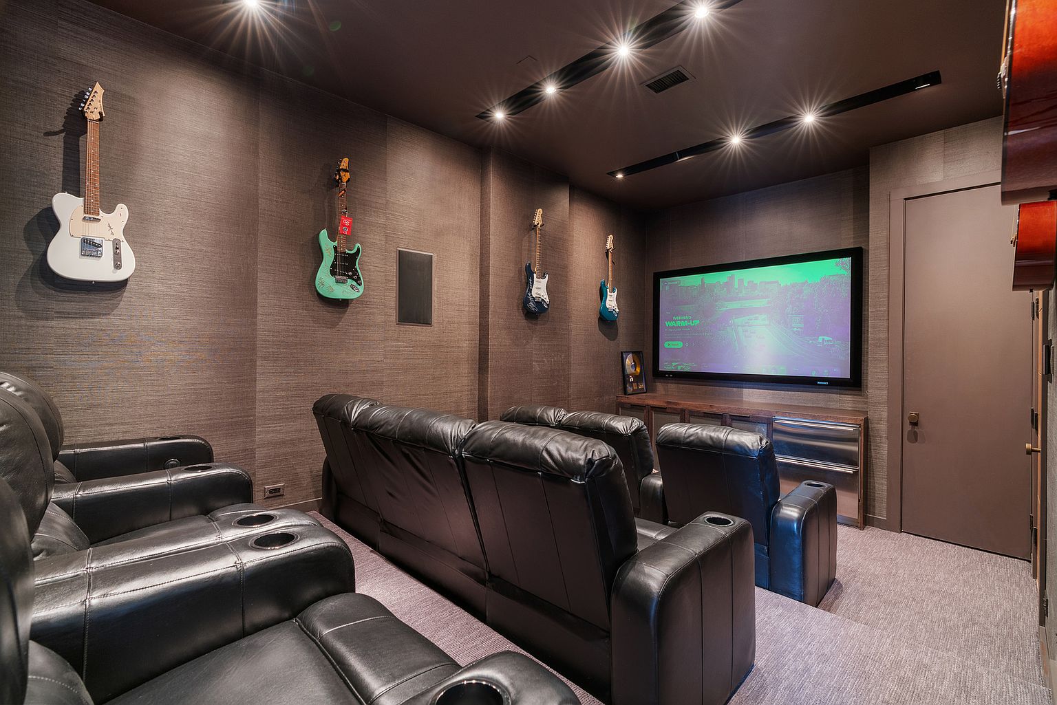 This dedicated home theater room features dark, textured wall coverings and a tiered seating arrangement with plush black leather recliners. The room is decorated with several electric guitars mounted on the walls, creating a personalized entertainment space. A large flat-screen television is mounted above a low-profile media console, providing a cinematic viewing experience in a cozy, enclosed environment.