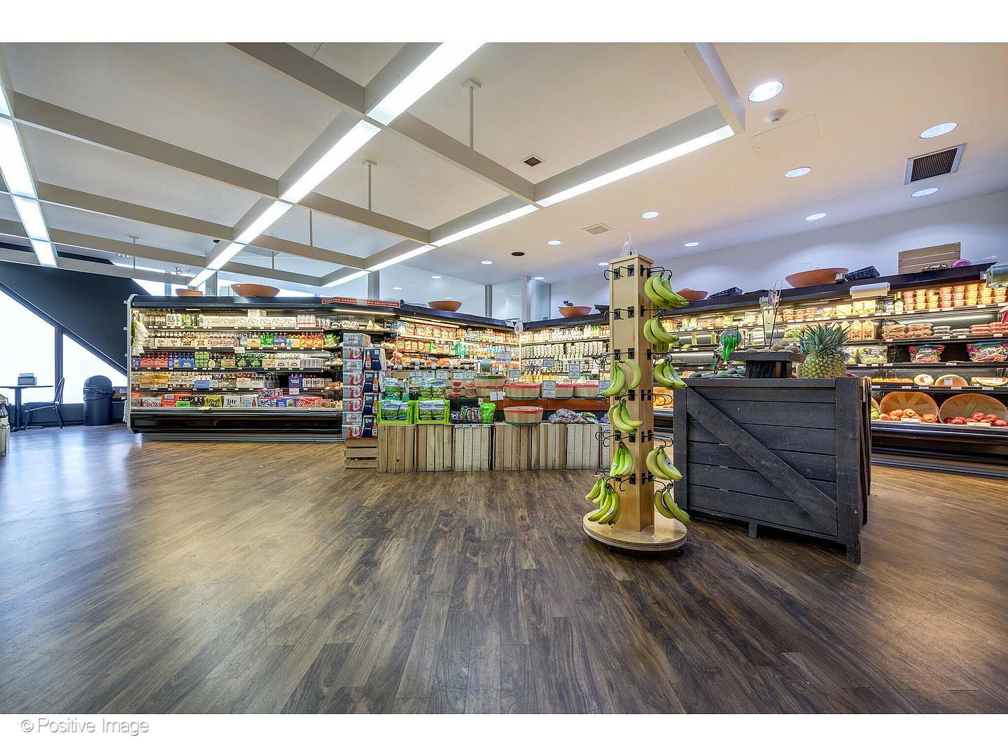 This image shows the interior of a commercial grocery store or market, featuring long refrigerated display cases stocked with various beverages and produce. A central wooden display stand holds fresh bananas, while wooden crates are used to organize fresh vegetables in the foreground. The space is illuminated by bright, linear overhead lighting, creating a clean and organized retail environment.