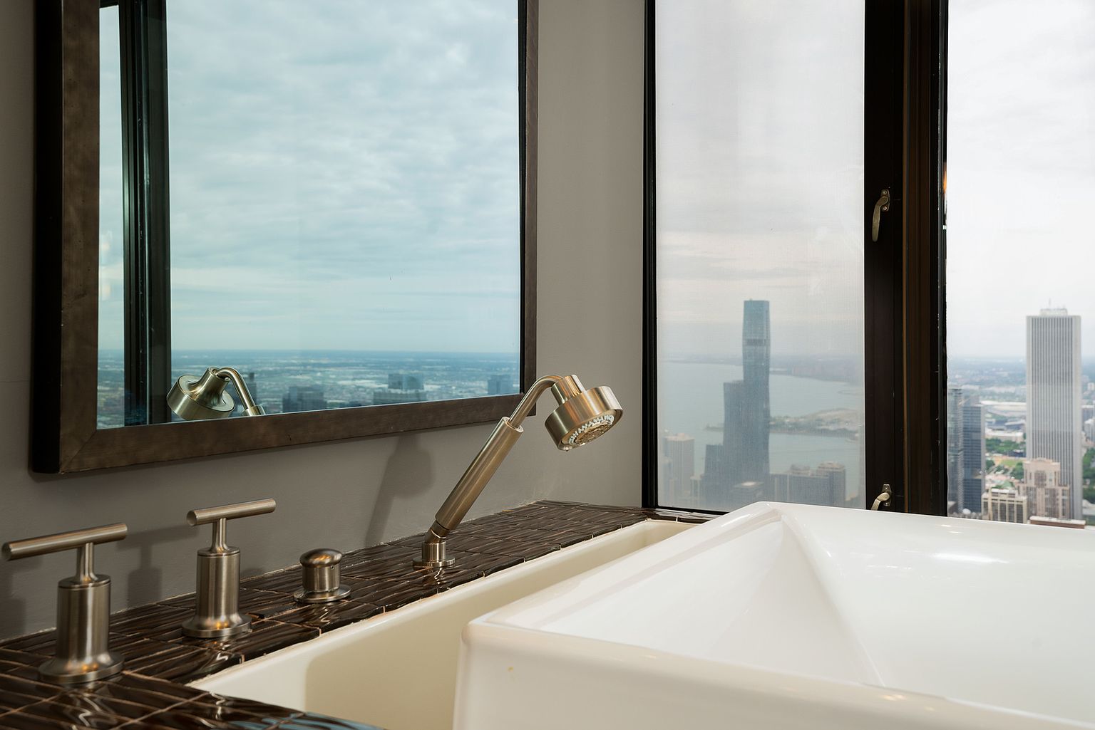 This high-rise bathroom features a deep soaking tub set against a backdrop of expansive, tall windows that offer a dramatic, panoramic view of the city skyline and waterfront. The tub is accented by a sleek, metallic handheld shower fixture and faucet set mounted on a dark, glossy tiled deck. The perspective captures the juxtaposition of luxurious indoor bathing amenities with the vast, urban landscape outside.