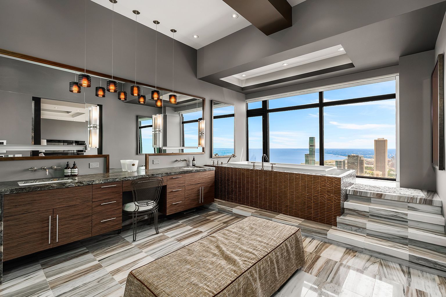 This luxurious primary bathroom features a long vanity with dark wood cabinetry and a stone countertop, complemented by a central makeup station and a matching upholstered bench. A deep soaking tub is set against a backdrop of large windows that provide expansive city and water views, with the tub area elevated by a set of steps clad in patterned stone. The space is illuminated by a series of modern pendant lights and recessed ceiling lighting, creating a sophisticated and high-end atmosphere.