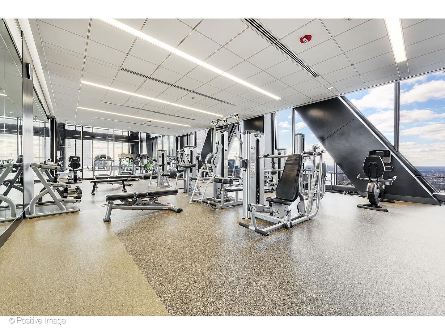 This professional-grade fitness center features a variety of strength training equipment, including weight machines and a flat bench, arranged on a durable, speckled rubber floor. The space is defined by expansive floor-to-ceiling windows that offer a panoramic view of the city skyline, creating an airy and motivating atmosphere. The room is illuminated by long, recessed linear lighting fixtures set into a clean, white drop ceiling.