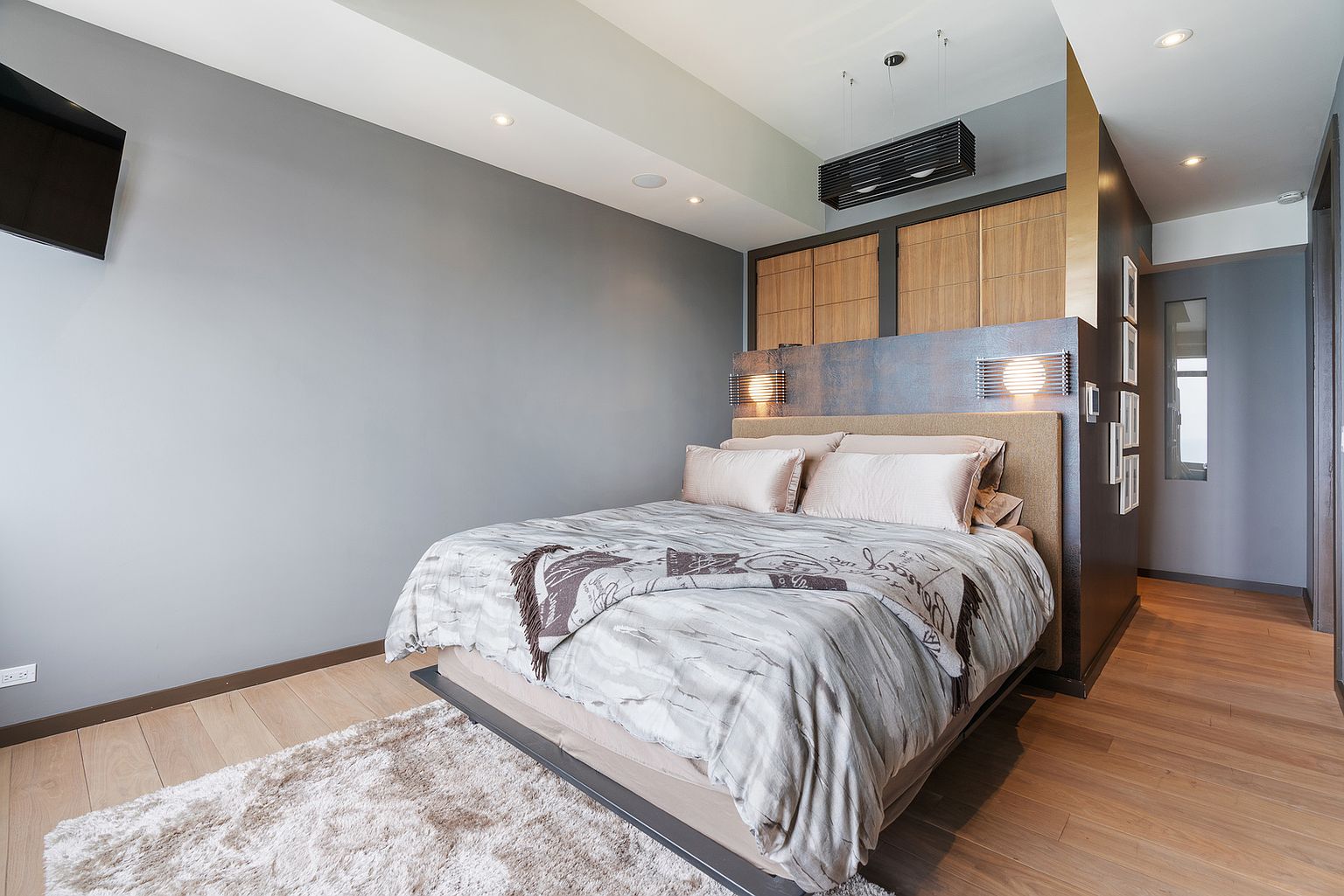 This contemporary primary bedroom features a platform bed centered against a dark, textured accent wall with integrated bedside lighting. Above the headboard, light wood cabinetry provides built-in storage, while a plush, textured rug sits atop the light wood flooring. The room is illuminated by a modern, linear pendant light and recessed ceiling lights, creating a sophisticated and cozy atmosphere.