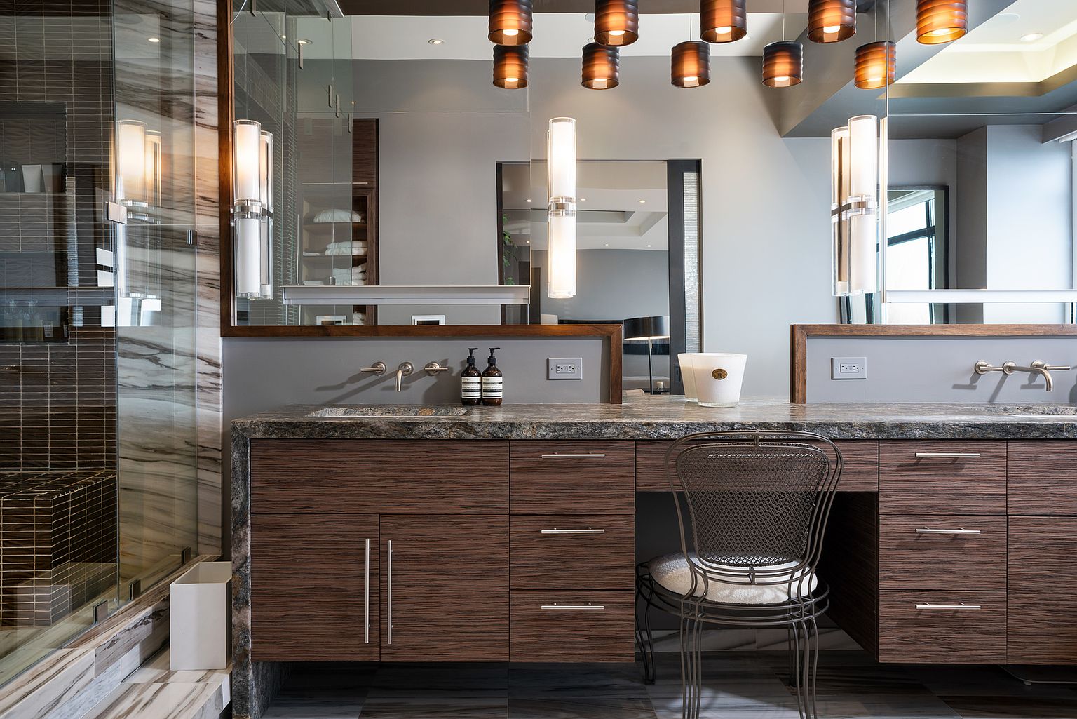This sophisticated primary bathroom features a dual-sink vanity with dark wood-toned cabinetry and a polished stone countertop. The space is accented by modern wall-mounted light fixtures, a metal mesh chair at the vanity station, and a glass-enclosed shower area with tiled walls. The perspective is a straight-on, eye-level shot that highlights the clean lines and contemporary design of the bathroom.