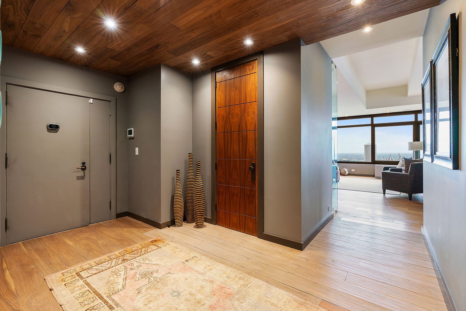 This sophisticated entryway features a warm wood-paneled ceiling with recessed lighting and light-toned wood flooring. A patterned area rug anchors the space, which leads toward a bright living area visible through a glass partition. The hallway is accented by a tall, modern wood door and a trio of decorative cylindrical floor vases, creating a clean, contemporary aesthetic.