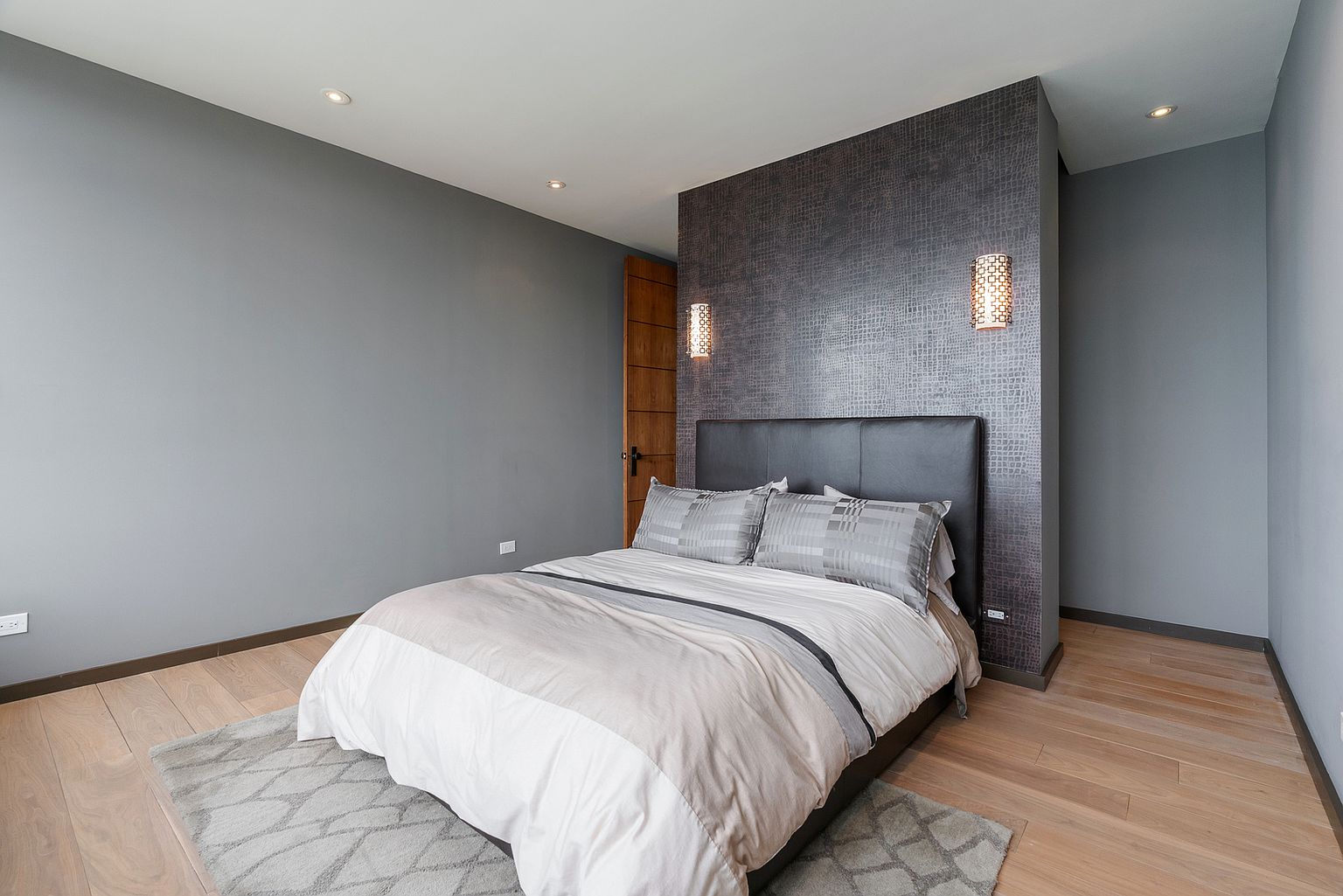 This modern primary bedroom features a dark, textured accent wall behind a leather-upholstered bed frame, creating a sophisticated focal point. The room is finished with light wood flooring, a neutral-toned area rug, and soft gray walls, complemented by two wall-mounted sconces. The perspective is a wide-angle shot from the foot of the bed, showcasing the clean, minimalist aesthetic and the warm, inviting atmosphere.