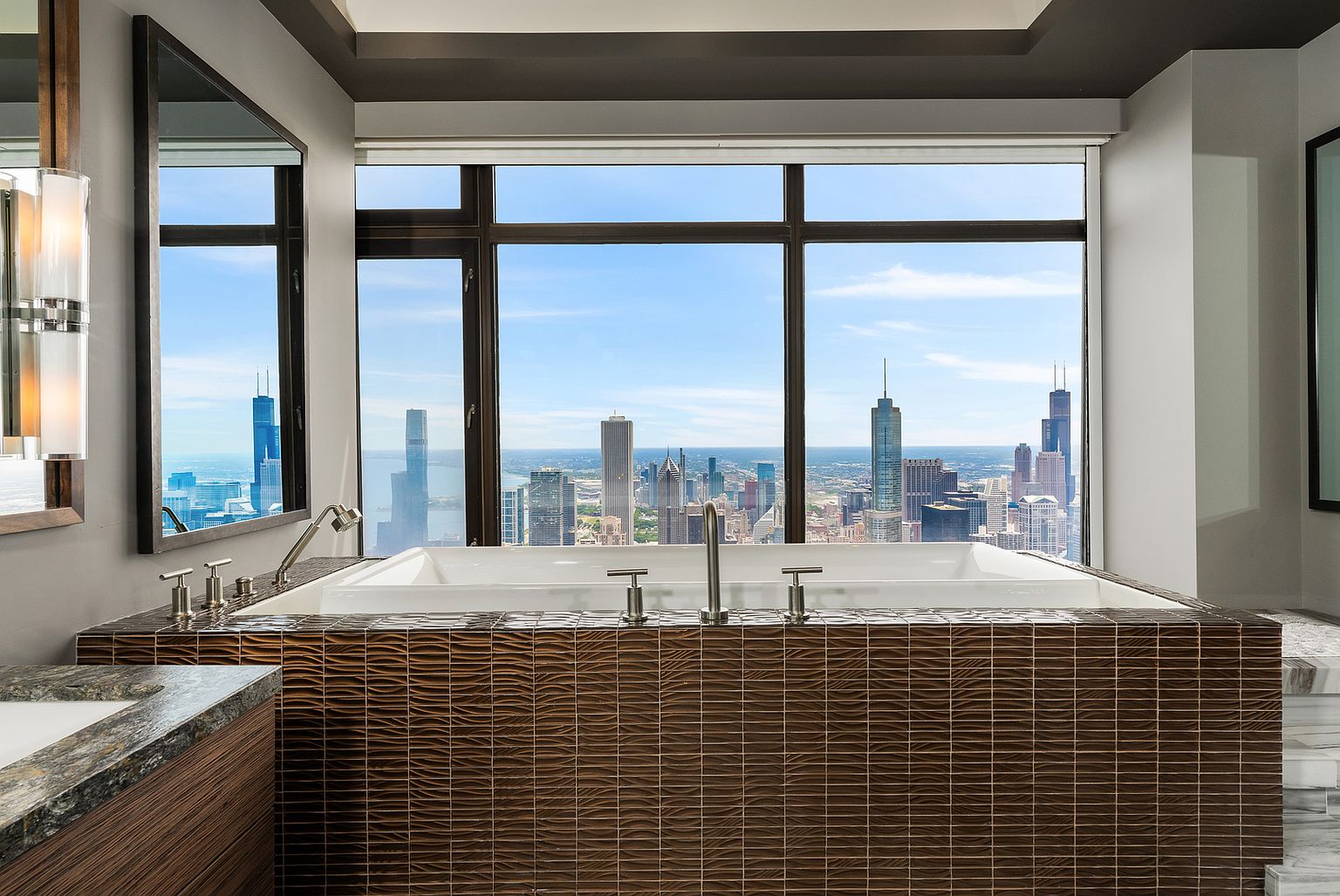 This luxurious primary bathroom features a deep soaking tub encased in textured, dark-toned horizontal tile, positioned directly in front of expansive windows that offer a panoramic view of the city skyline. To the left, a large mirror reflects the urban landscape, while modern chrome fixtures and a sleek vanity with a stone countertop complete the sophisticated aesthetic. The perspective is centered on the tub, emphasizing the dramatic contrast between the intimate, dark-tiled interior and the bright, open cityscape beyond.