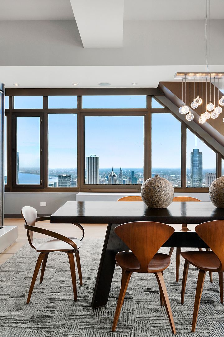 This sophisticated dining area features a dark wood table surrounded by mid-century modern style chairs on a textured gray area rug. A striking multi-pendant light fixture hangs above, while large windows provide an expansive, panoramic view of the city skyline and waterfront. The space is defined by its clean lines, neutral color palette, and a sense of elevated urban living.