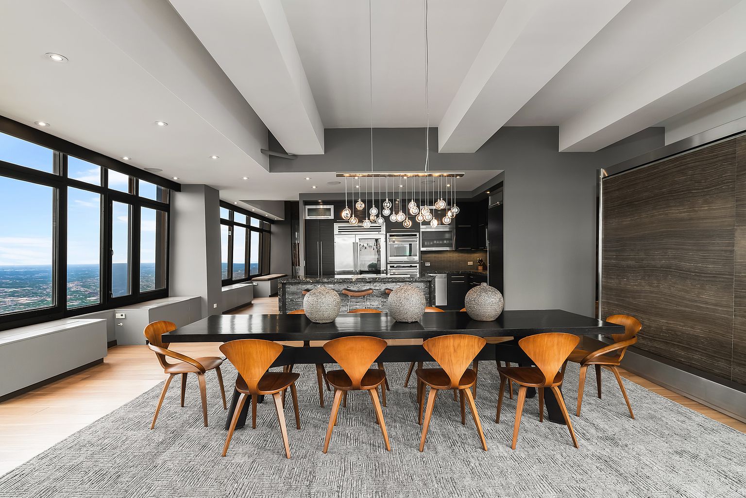 This sophisticated dining area features a long, dark table surrounded by eight mid-century modern style wooden chairs, all set upon a textured grey area rug. The space is illuminated by a striking multi-globe pendant light fixture and benefits from expansive windows that offer a panoramic view of the city skyline. In the background, a sleek, open-concept kitchen with dark cabinetry and a stone-topped island completes the contemporary, high-rise aesthetic.