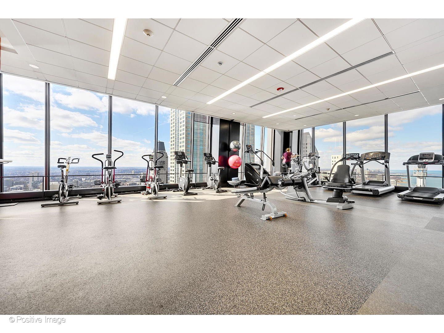 This expansive, modern home gym features a wide array of professional-grade exercise equipment, including stationary bikes, treadmills, and a weight bench. The space is defined by its impressive floor-to-ceiling windows that offer panoramic city views, creating an airy and inspiring workout environment. The room is finished with neutral, durable flooring and a clean, drop-ceiling design with integrated lighting.