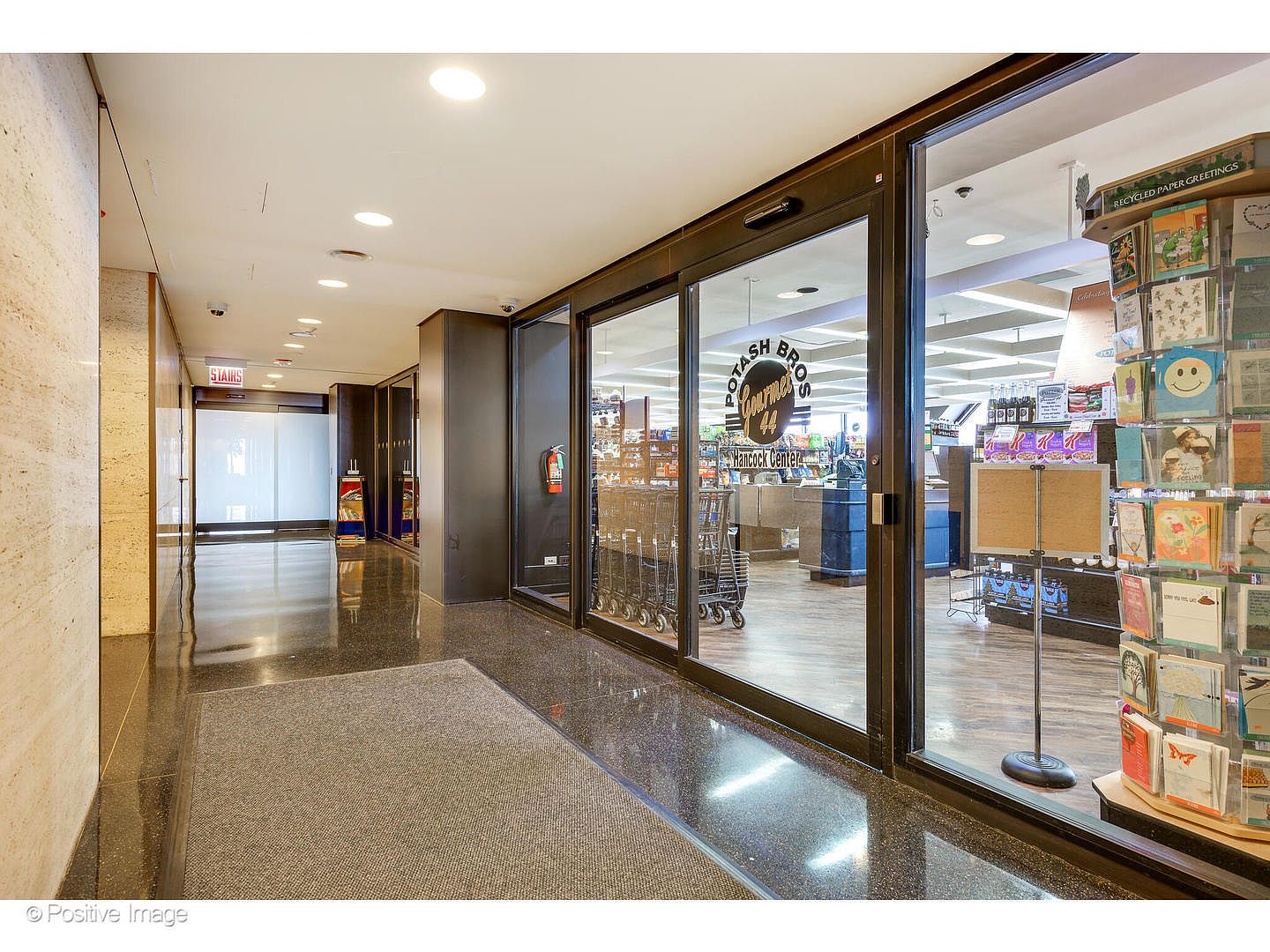 This image captures a polished, commercial-style hallway leading toward a glass-fronted grocery store entrance labeled 'Potash Bros Gourmet'. The space features a dark, reflective stone floor, recessed ceiling lighting, and a prominent display rack filled with greeting cards on the right. The perspective is a straight-on view down the corridor, emphasizing the clean, professional transition between the building's common area and the retail space.