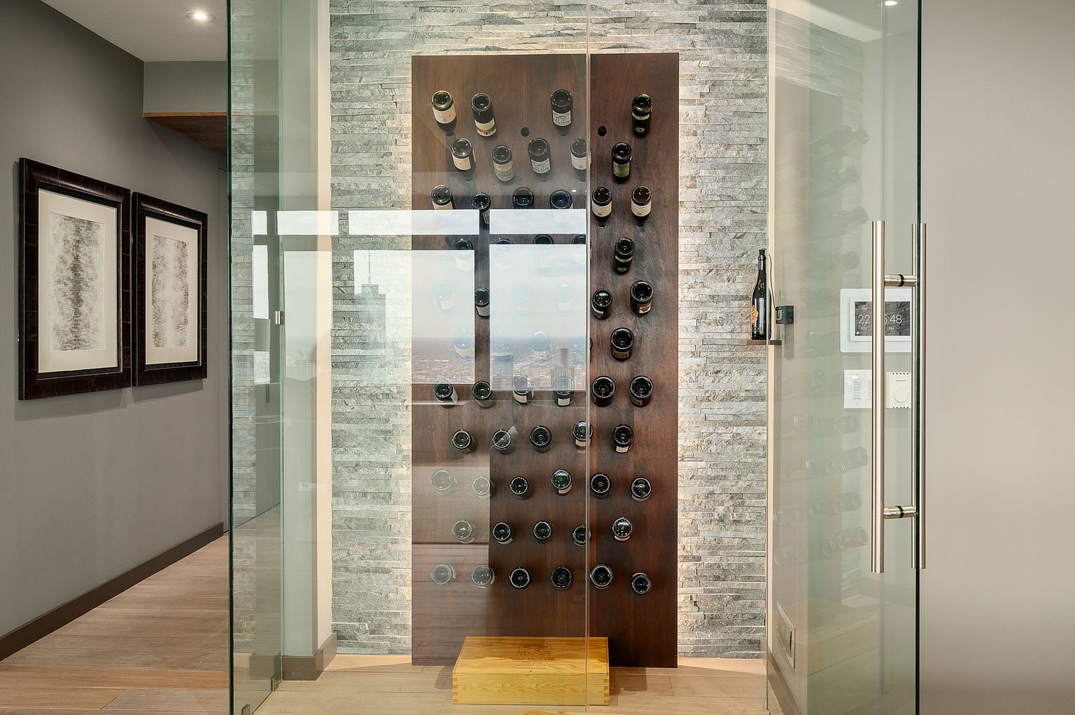 This sophisticated wine cellar features a glass-enclosed display area with a dark wood panel wall mounted with individual bottle racks. The surrounding walls are finished in textured, light-toned stacked stone, creating a modern and elegant aesthetic. A wooden crate sits on the floor at the base of the display, and the perspective looks directly through the glass door into the climate-controlled storage space.