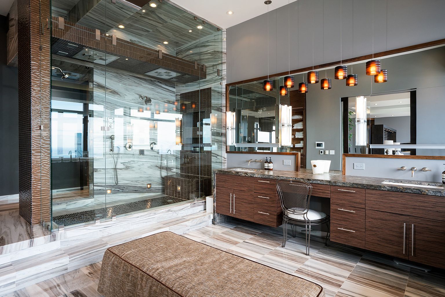 This luxurious primary bathroom features a large, glass-enclosed walk-in shower with patterned stone walls and a built-in bench. A long vanity with dark wood cabinetry and a stone countertop provides ample space, complemented by a vanity chair and a series of modern pendant lights hanging above the mirror. The room is finished with large-format stone floor tiles and a central upholstered bench, creating a sophisticated and high-end atmosphere.