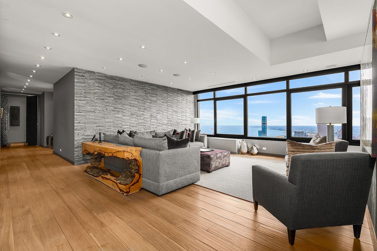 This spacious, modern living room features a prominent stacked-stone feature wall and light wood flooring that extends into a hallway. A large gray sectional sofa is accented by a unique, raw-edge wood console table, while expansive windows provide panoramic city and water views. The room is illuminated by recessed lighting and includes a comfortable armchair, creating an inviting and sophisticated atmosphere.