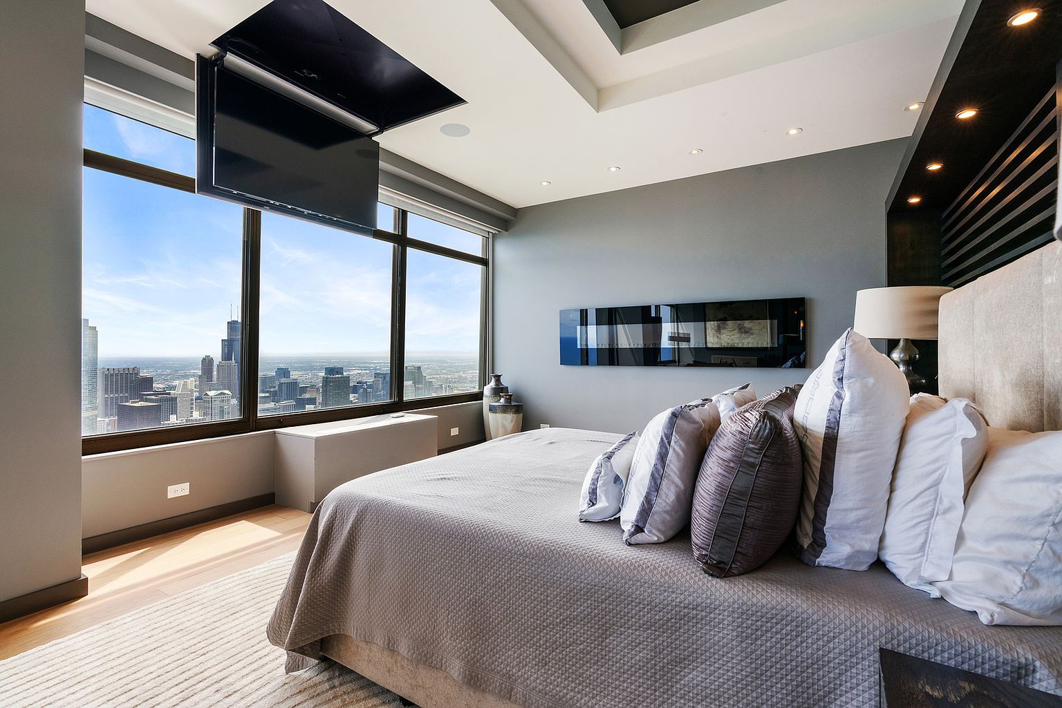 This sophisticated primary bedroom features a neutral color palette and a plush bed adorned with layered pillows. A motorized television descends from the ceiling, positioned perfectly for viewing from the bed, while expansive windows provide a dramatic, unobstructed panoramic view of the city skyline. The room is finished with recessed lighting and a sleek, modern aesthetic that emphasizes the high-rise urban setting.