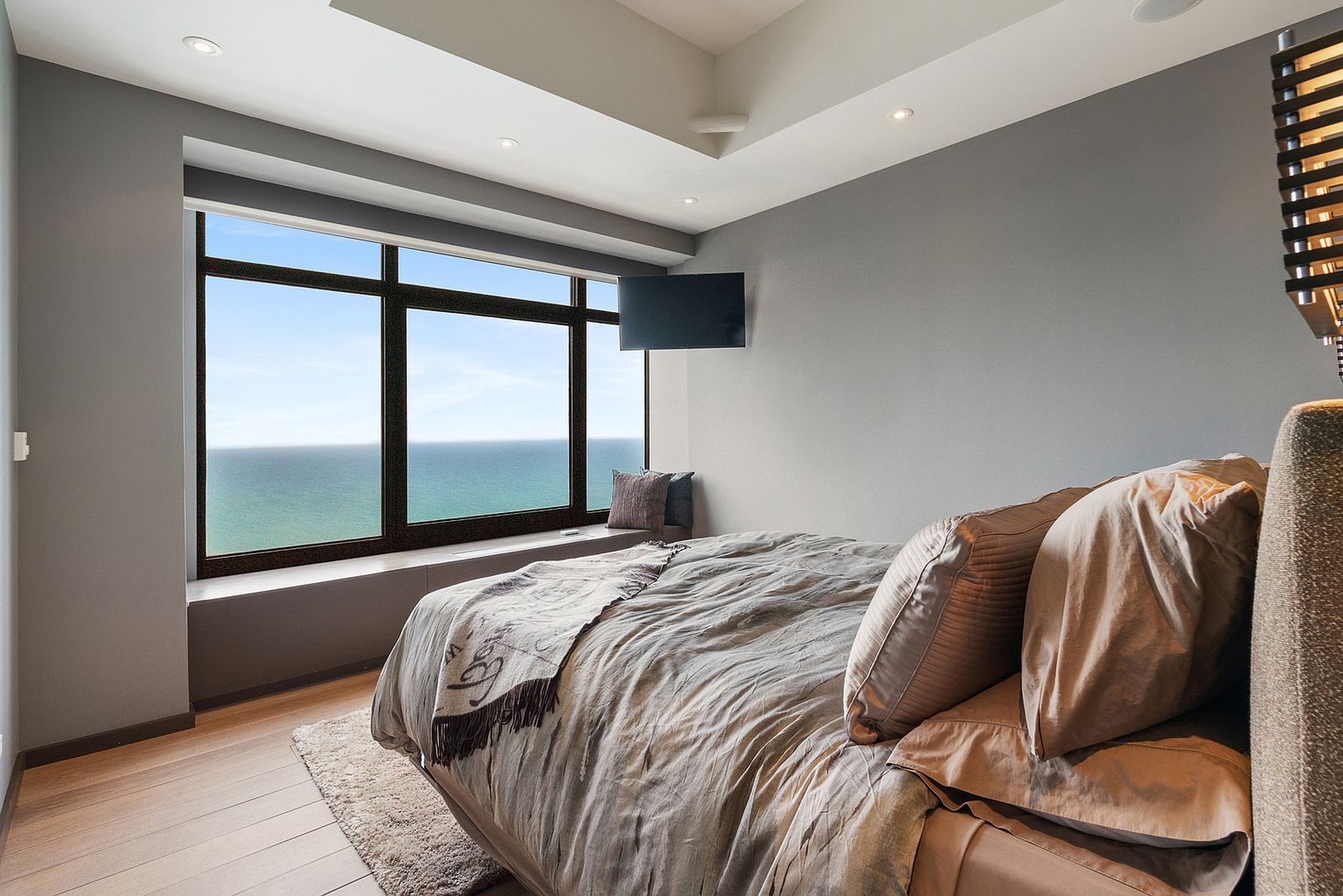 This serene primary bedroom features a neutral color palette and a large window that frames a panoramic view of the ocean. The room is furnished with a plush bed dressed in soft linens and a textured throw, positioned to take advantage of the natural light. A wall-mounted television and a built-in window seat complete the space, creating a calm and sophisticated retreat.
