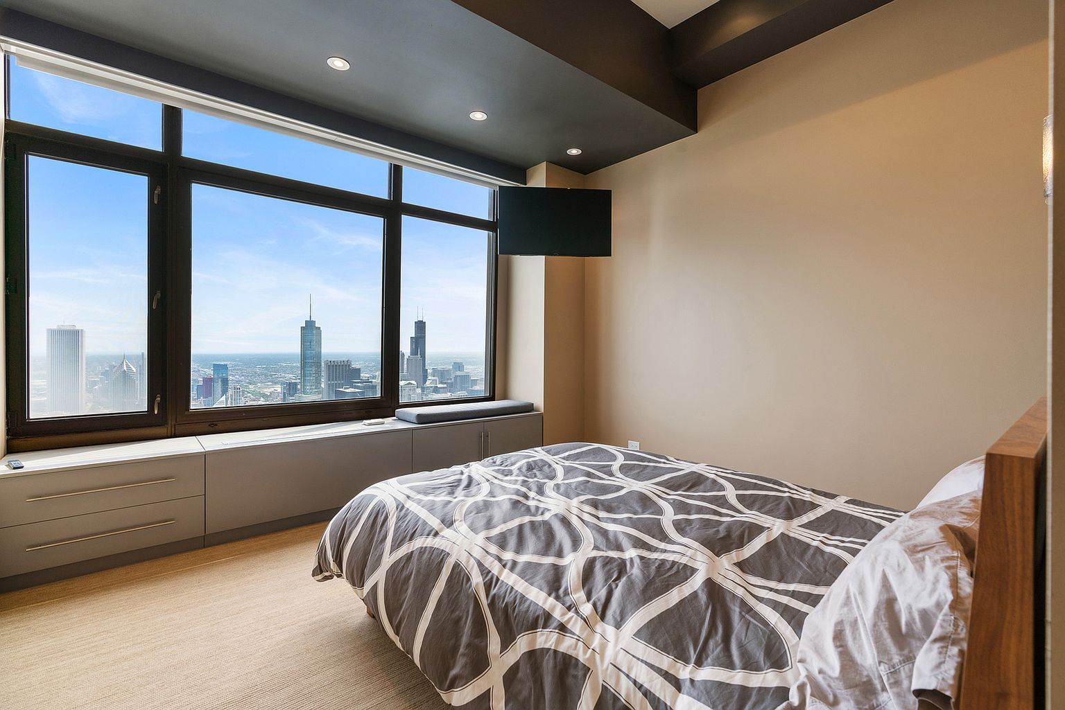 This bedroom features a modern aesthetic with a bed dressed in a geometric patterned duvet and a wooden headboard. A large window wall provides an expansive, elevated view of the city skyline, complemented by built-in cabinetry and a window seat below. The room is finished with neutral-toned walls and a dark ceiling accent, creating a sleek and contemporary urban living space.