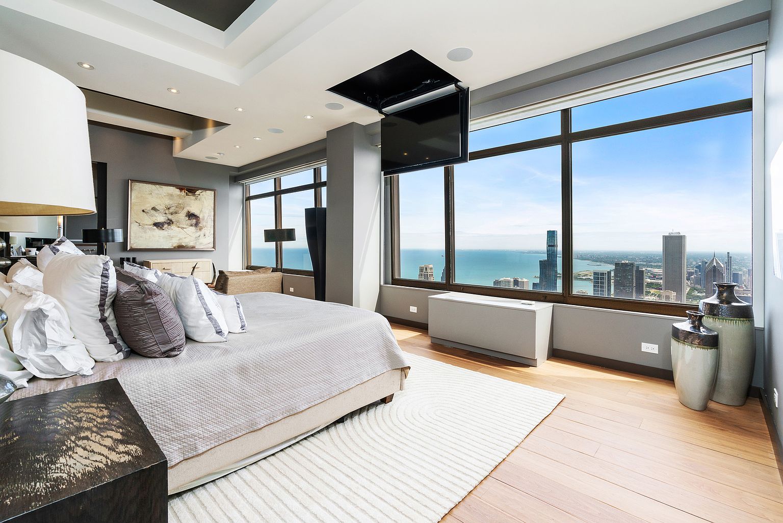 This luxurious primary bedroom features a plush bed centered in the room, complemented by a textured area rug and a dark wood nightstand. Large windows provide expansive, unobstructed views of the city skyline and the lake, creating a bright and airy atmosphere. A television is mounted on a motorized lift system integrated into the ceiling, adding a sophisticated, modern touch to the space.