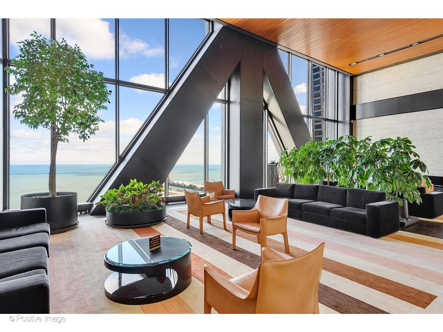 This sophisticated lounge area features expansive floor-to-ceiling windows that offer panoramic views of the water and city skyline. The space is furnished with a long black sofa and several tan leather armchairs arranged on a striped area rug, complemented by potted greenery and a circular glass-topped coffee table. The modern architectural design includes a striking angled structural beam and a warm wood-paneled ceiling, creating an airy and upscale atmosphere.