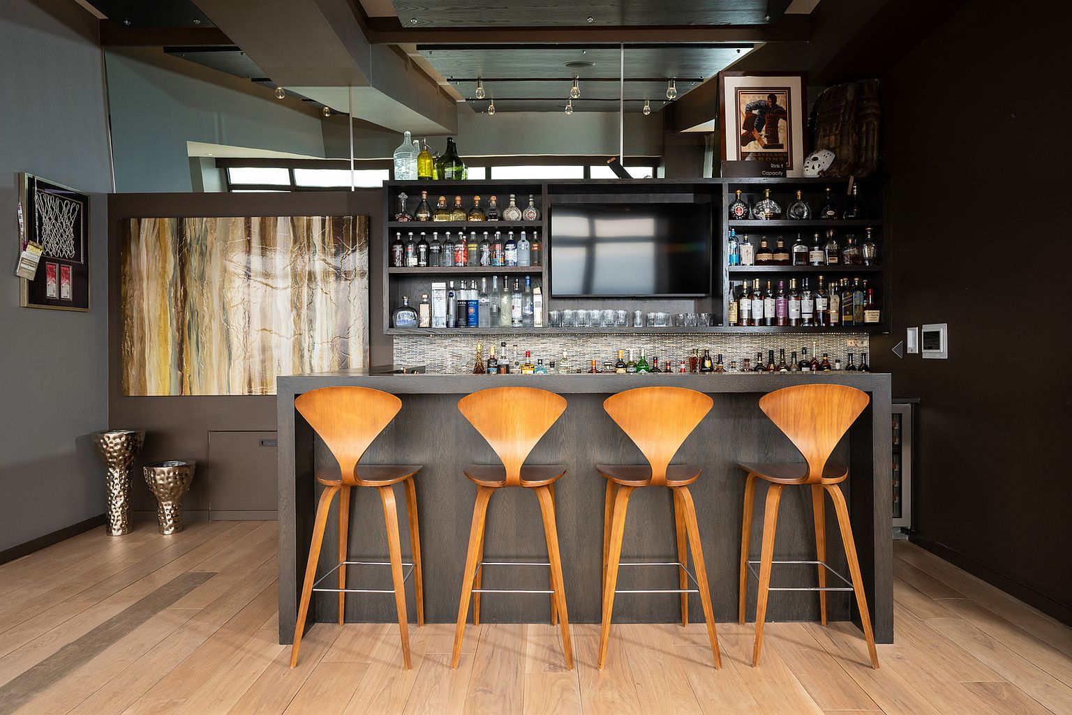 This home bar area features a dark, modern bar counter with four curved wooden stools arranged in a row. Behind the bar, a built-in shelving unit displays an extensive collection of liquor bottles and glassware, centered around a wall-mounted television. The space is accented by a large abstract art piece on the left wall and two metallic decorative vessels on the floor, creating a sophisticated and masculine entertainment atmosphere.