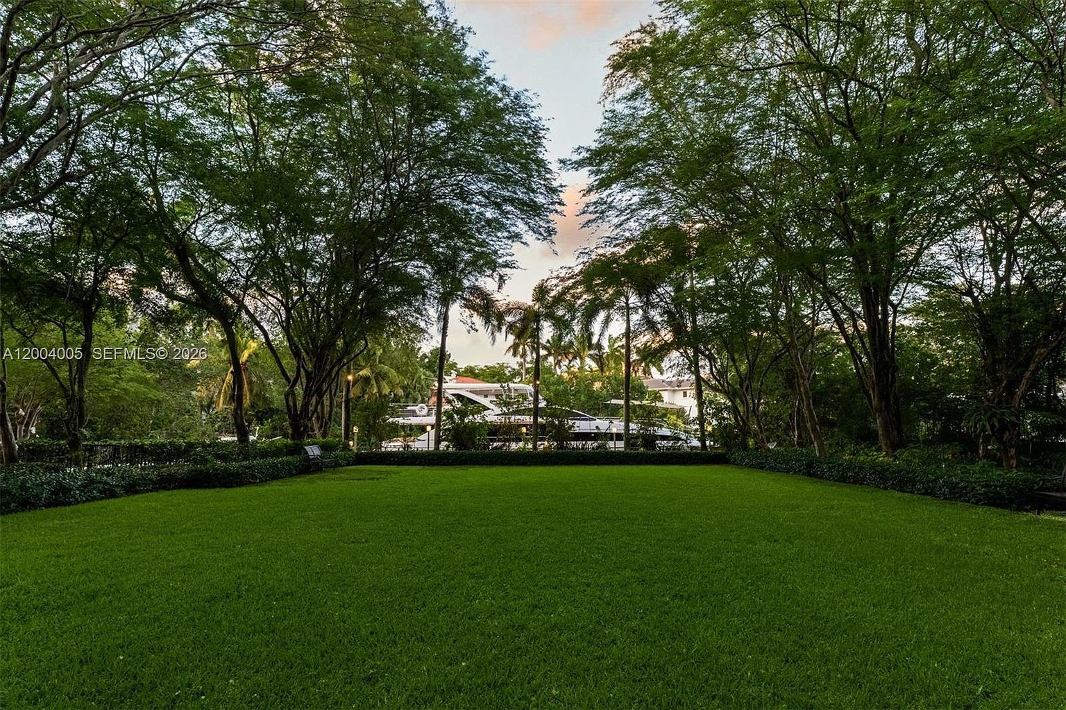 A expansive, well-manicured green lawn serves as the foreground, framed by mature, leafy trees that create a natural canopy. In the background, a large white yacht is visible docked along the water, partially obscured by the lush foliage. The perspective is a low-angle, eye-level shot looking across the lawn toward the water, evoking a sense of privacy and serene waterfront living.