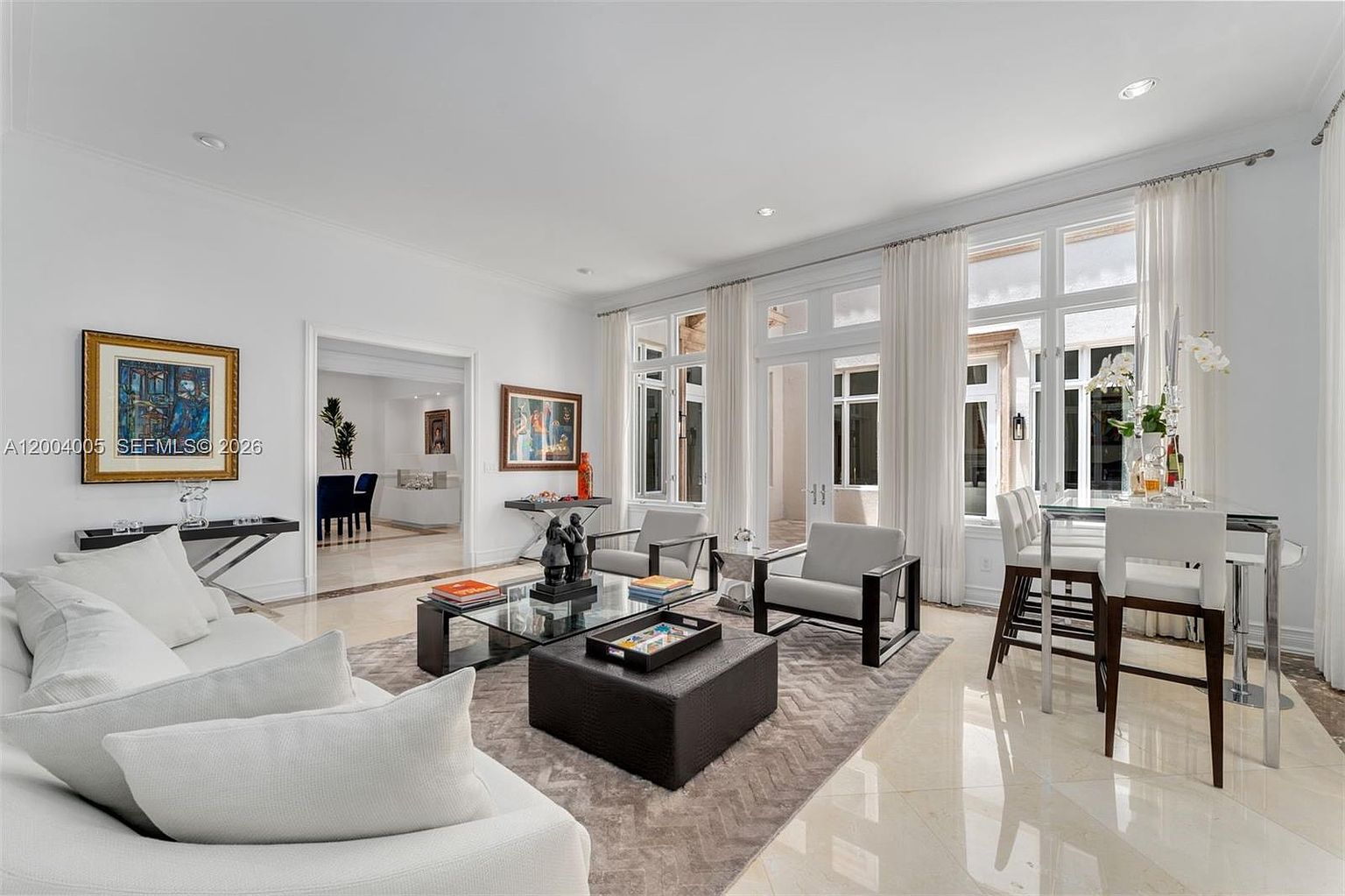 This bright and airy living room features a neutral color palette with white sofas, a dark leather ottoman, and two modern armchairs arranged on a textured area rug. Large windows with sheer curtains allow ample natural light to fill the space, which also includes a glass-topped dining table and framed artwork on the walls. The polished stone flooring and open floor plan create an elegant and sophisticated atmosphere suitable for entertaining.