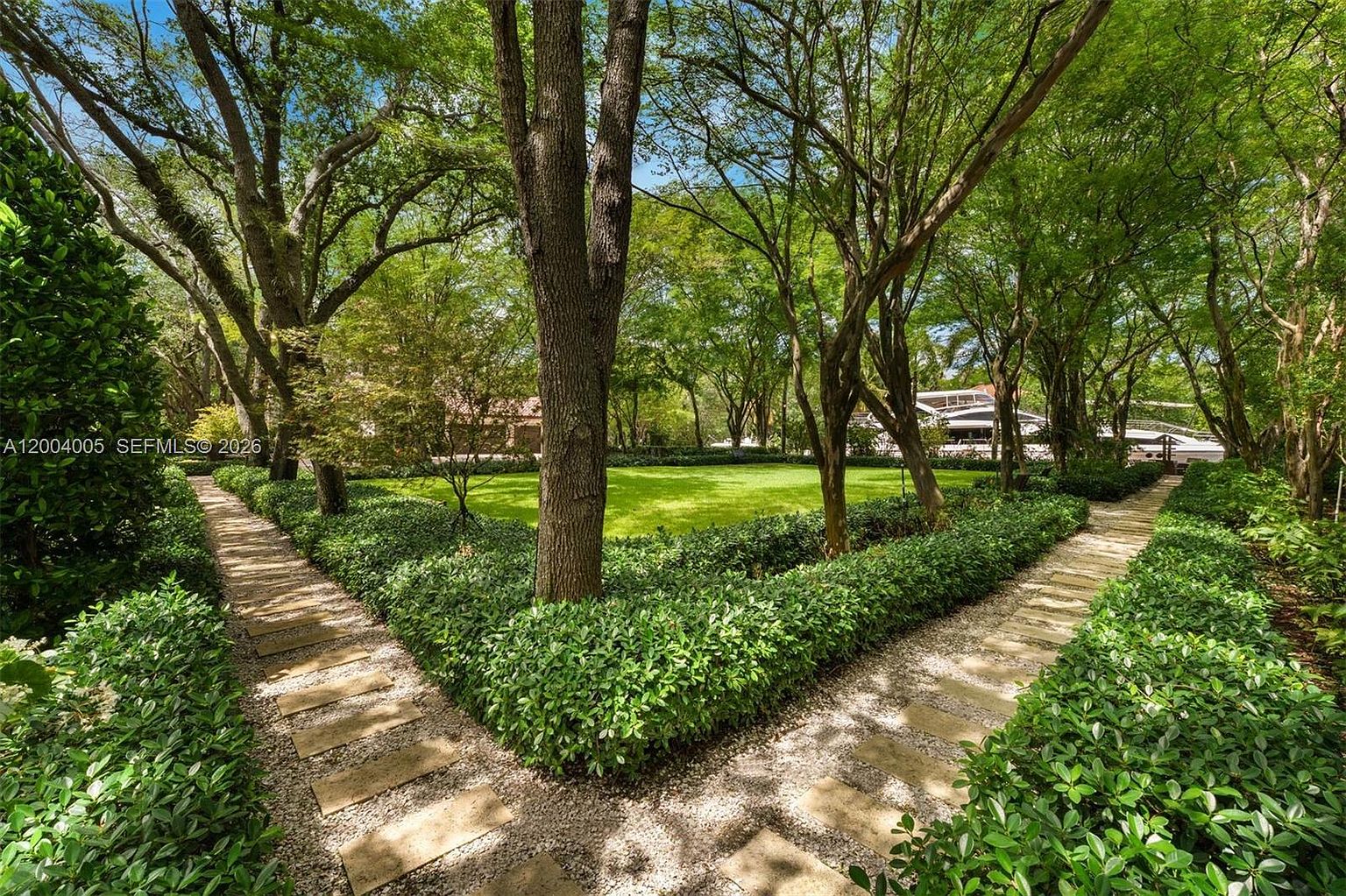 This lush, park-like garden features a manicured green lawn framed by mature, leafy trees that create a canopy overhead. Two stone-paver pathways lined with neatly trimmed hedges converge toward the center of the yard, offering a symmetrical and inviting perspective. The scene evokes a serene, private estate atmosphere with dappled sunlight filtering through the dense foliage.