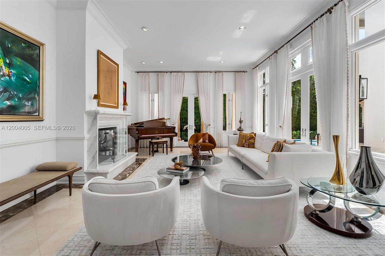 This elegant living room features a bright, airy aesthetic with white walls and large windows draped in sheer curtains that allow ample natural light. The space is anchored by a central seating area with a white sofa, two matching armchairs, and a pair of circular coffee tables, all set upon a textured area rug. A grand piano sits near the tall stone-clad fireplace, while a long bench and decorative accents complete the sophisticated, open-plan layout.