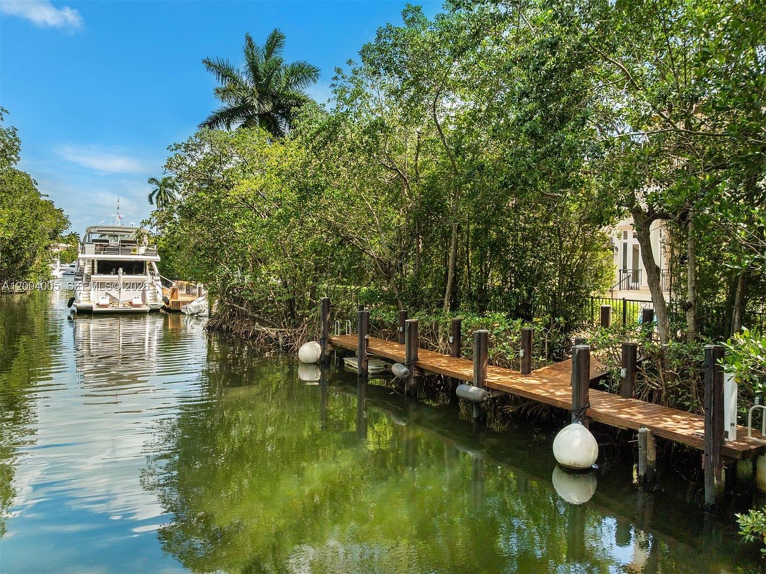This view captures a serene waterfront setting featuring a wooden dock extending into a calm canal lined with lush, mature trees. A large yacht is moored in the distance, emphasizing the property's accessibility for boating enthusiasts. The perspective is from the water looking toward the property, highlighting the private dock and the natural, tranquil landscape.