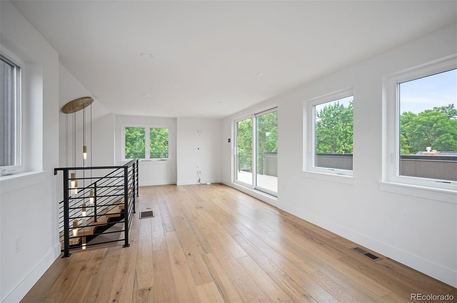 This is a bright and airy living room featuring light hardwood floors and white walls, creating a modern and spacious feel. Large windows and a sliding glass door provide ample natural light and views of the outdoors. A staircase with black metal railings adds a contemporary architectural element to the space.