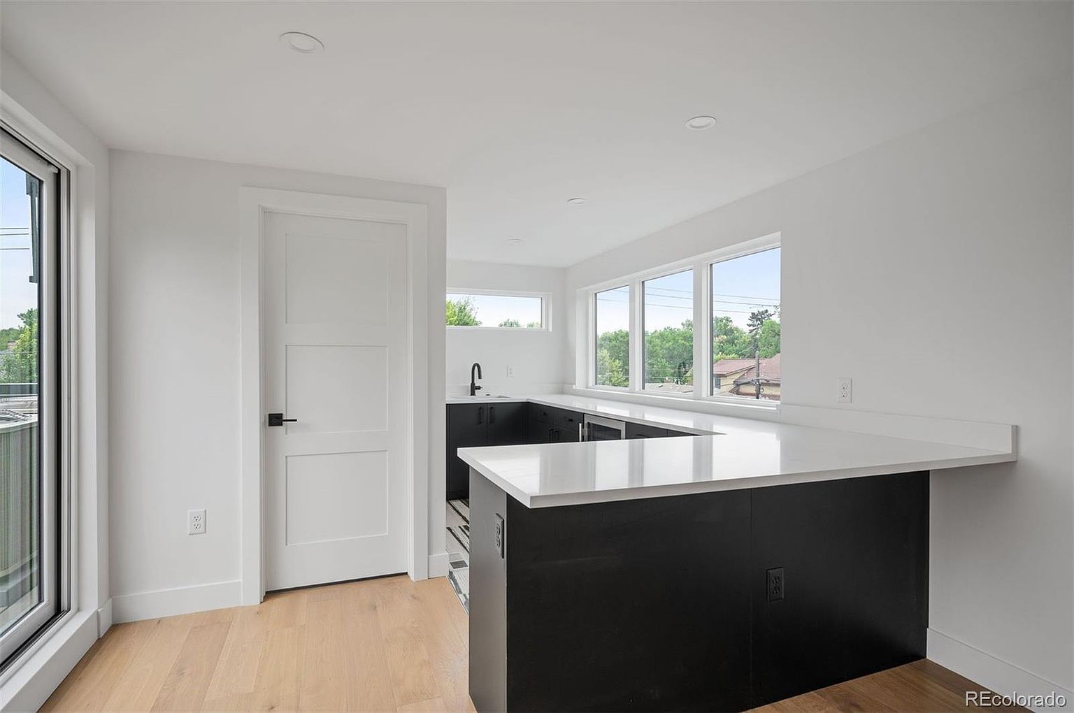 This is a bright and modern kitchen featuring a black and white color scheme. The kitchen island has a white countertop and black base, complemented by black cabinetry and fixtures. Large windows provide ample natural light, enhancing the clean and minimalist aesthetic of the space.