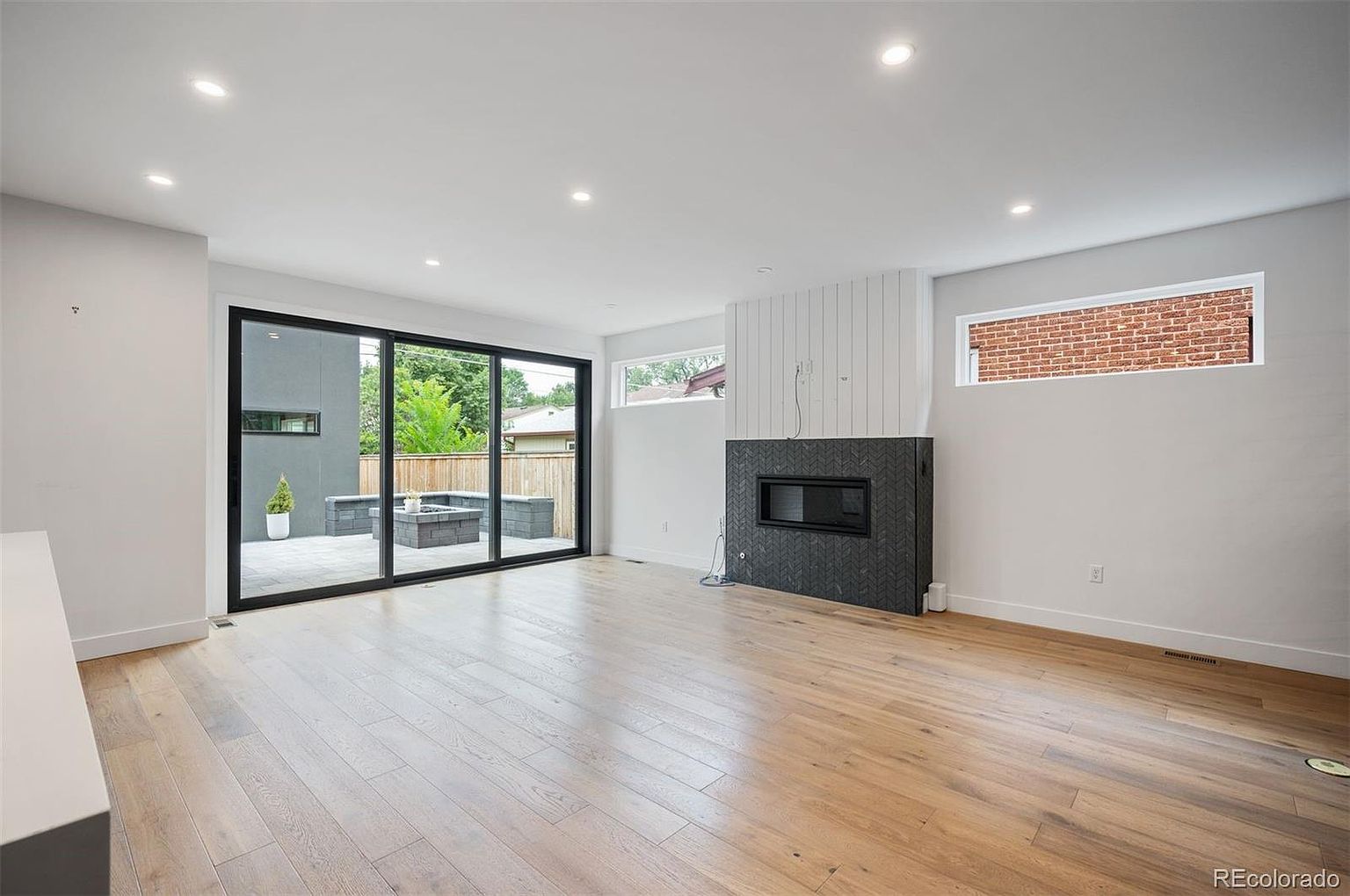 This is a bright and spacious living room featuring hardwood floors and recessed lighting. A modern fireplace with a dark herringbone tile surround adds a focal point, while a large sliding glass door provides access to an outdoor patio area. The room's neutral color palette and clean lines create a contemporary and inviting atmosphere.