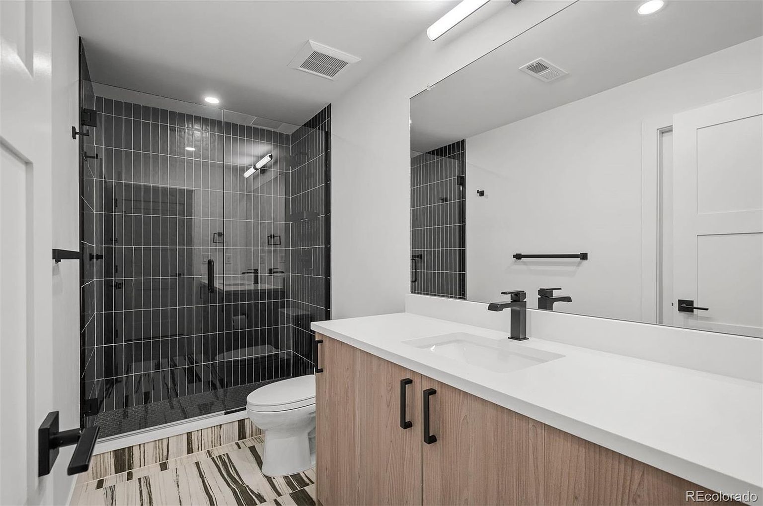 This is a modern bathroom featuring a walk-in shower with black-framed glass and dark gray vertical tiles. The vanity has a light wood finish with a white countertop and a black faucet. The flooring is a unique patterned tile, and a large mirror reflects the space, enhancing the sense of openness.