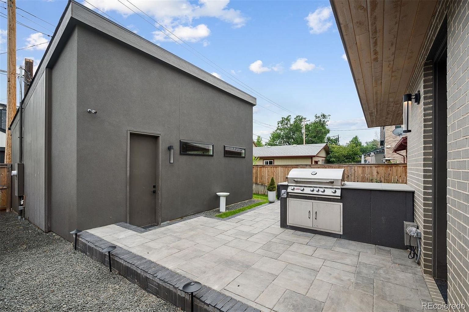 This image showcases a modern patio area, featuring a built-in outdoor kitchen with a stainless steel grill and dark cabinetry. The patio is paved with large, light-colored stones, and a dark gray building with a minimalist design stands adjacent to the space. The overall impression is clean, contemporary, and ideal for outdoor entertaining.
