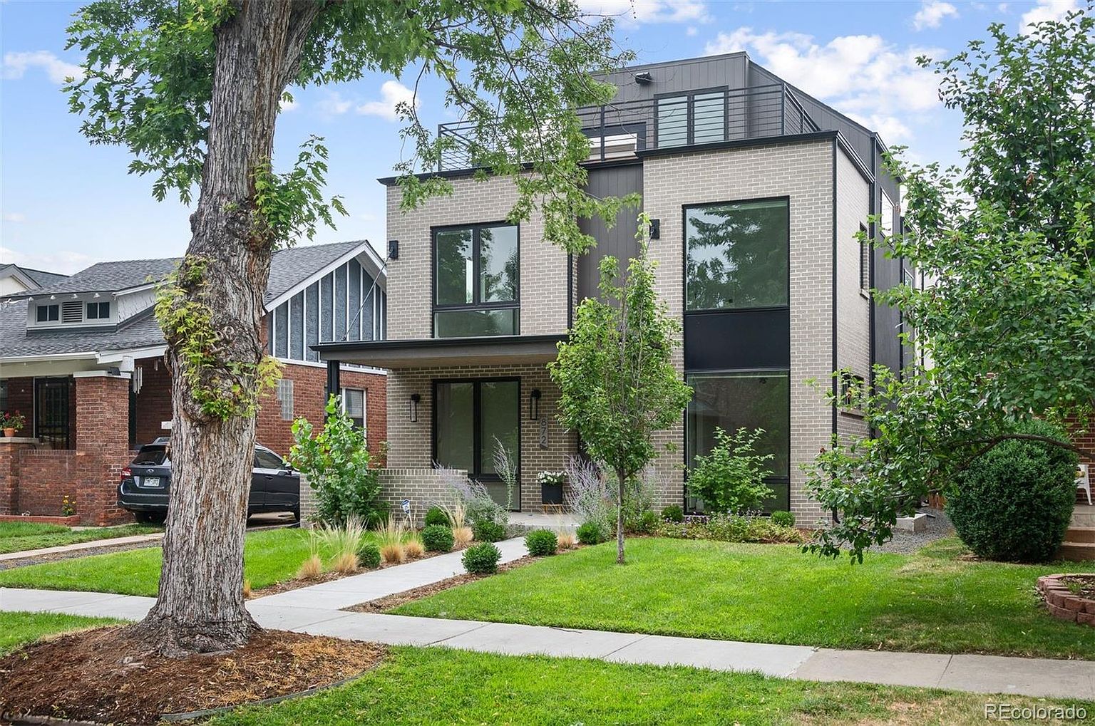 This is a front exterior view of a modern two-story home with a light brick facade and dark trim. The house features large windows, a small front yard with a well-maintained lawn, and a concrete walkway leading to the entrance. Mature trees frame the property, adding to its curb appeal.