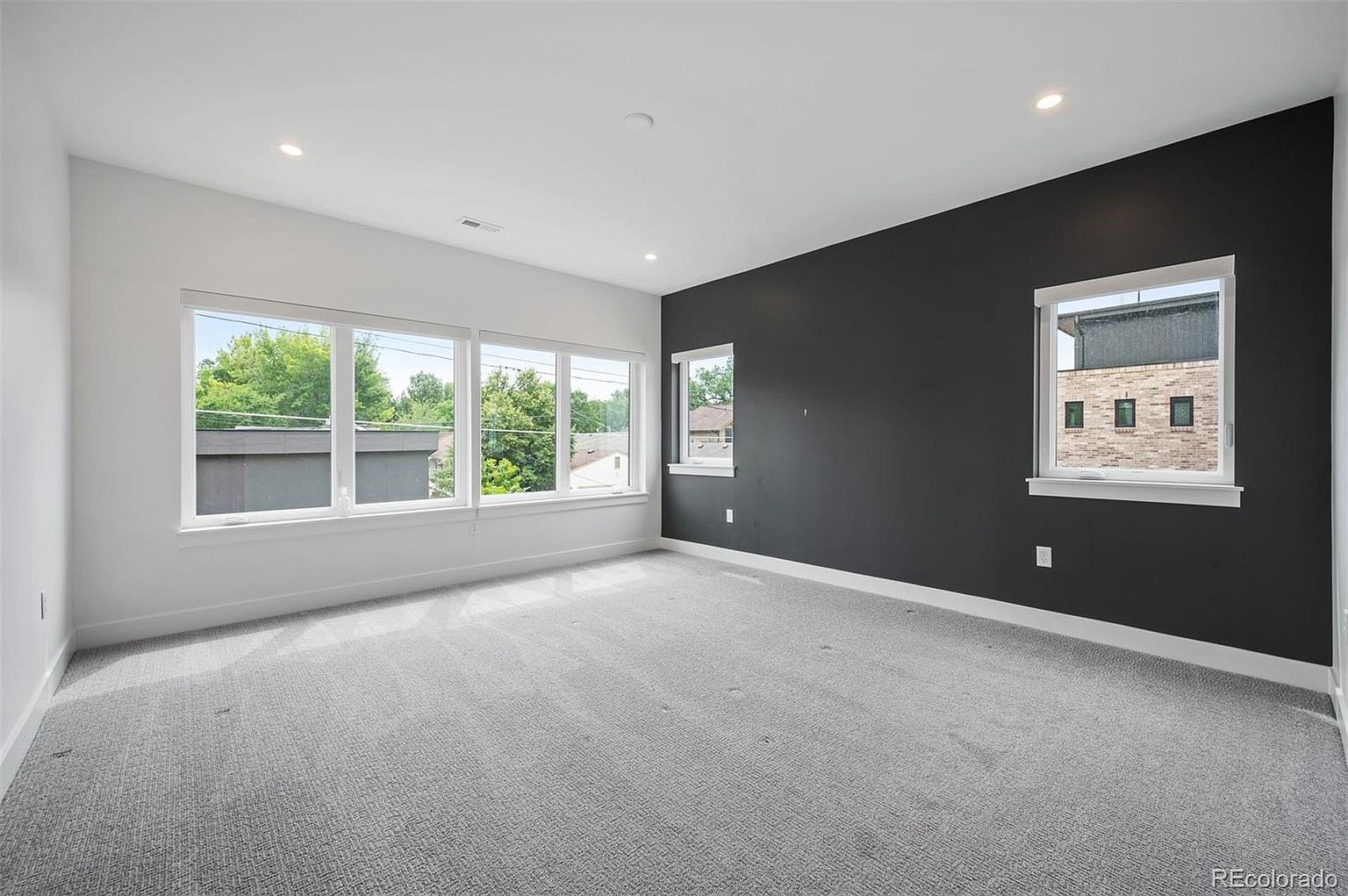 This is a spacious bedroom featuring a modern design with a contrasting color scheme. One wall is painted a dark gray, while the others are white, creating a striking visual effect. Large windows provide ample natural light, and the room is carpeted with a neutral gray carpet, contributing to a clean and contemporary aesthetic.