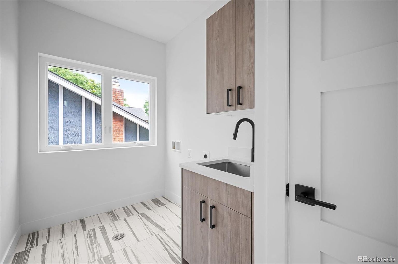 This is a bright and modern laundry room featuring a sleek vanity with a stainless steel sink and matte black faucet. The room has a window providing natural light and a view of the exterior. The flooring is a stylish tile with a linear pattern, and the cabinetry is a light wood tone with black hardware, creating a clean and contemporary aesthetic.
