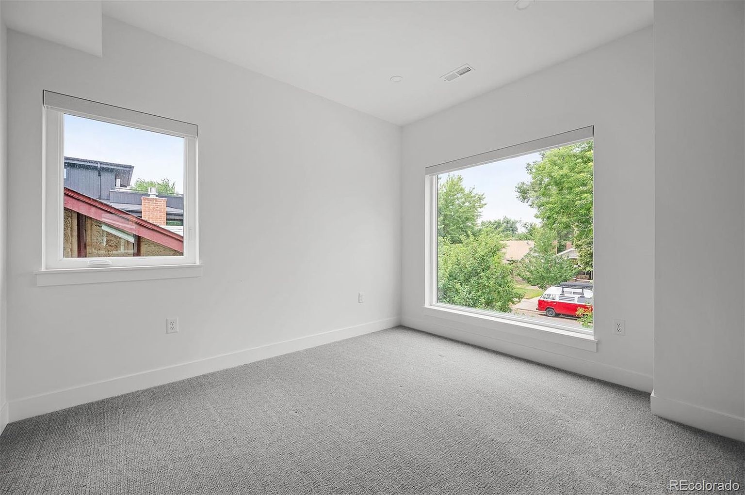This is a bright and airy bedroom with neutral gray carpet and white walls. Two large windows provide ample natural light and views of the outdoors. The room is clean and minimalist, offering a blank canvas for potential buyers to envision their own style.