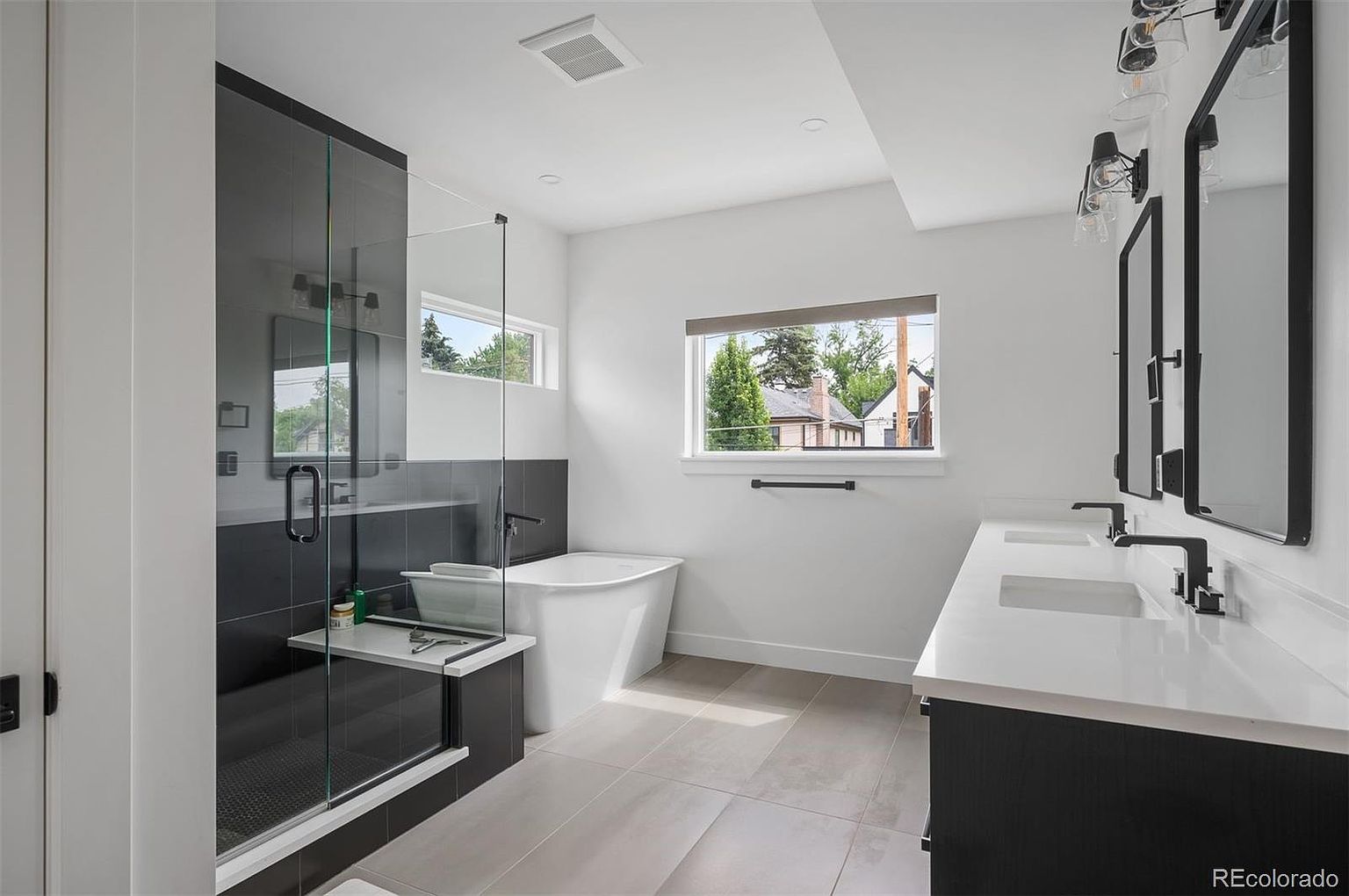 This is a modern primary bathroom featuring a glass-enclosed shower with dark gray tiling and a built-in bench. A freestanding white bathtub sits beneath a window, and a double vanity with black cabinets and white countertops is visible on the right. The overall aesthetic is clean, contemporary, and luxurious.