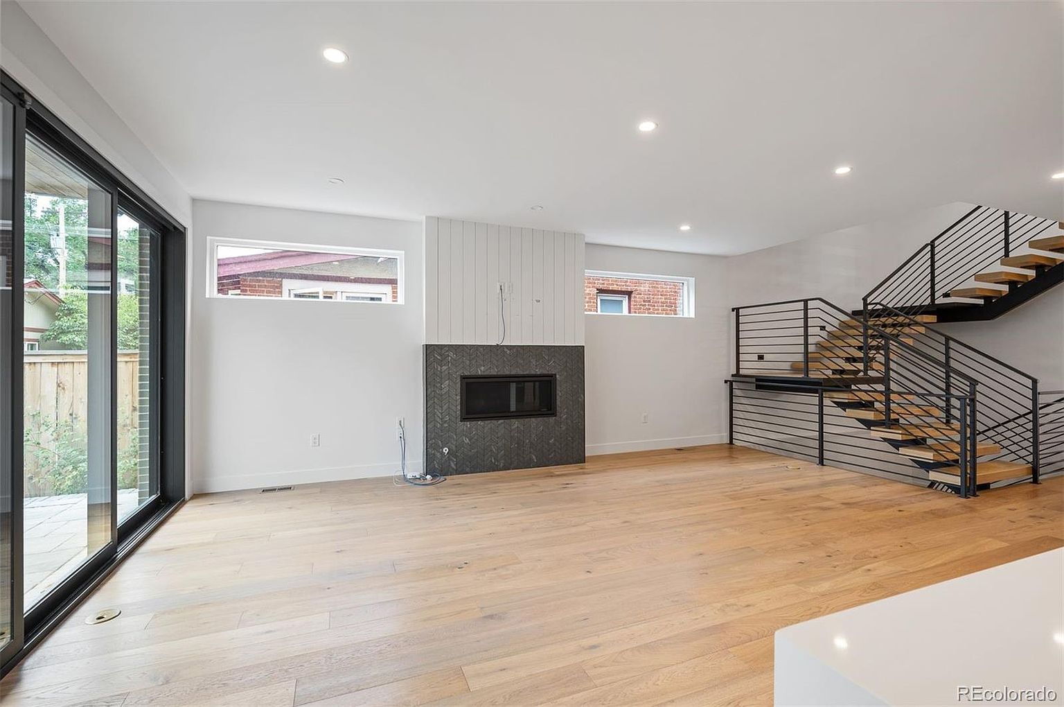 This is a bright and modern living room featuring light hardwood floors, a contemporary fireplace with a dark tile surround, and a staircase with metal railings and wooden steps. Large sliding glass doors provide ample natural light and access to the outside. The room has a minimalist aesthetic with clean lines and a neutral color palette.