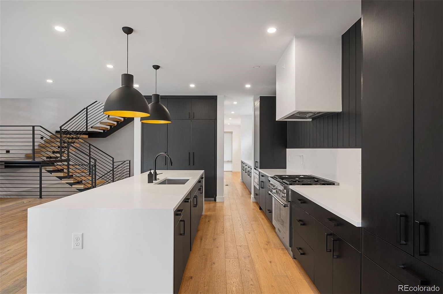 This is a modern kitchen with a long island featuring a white countertop and dark cabinetry. The kitchen also features stainless steel appliances, a white range hood, and dark cabinets along the wall. The hardwood floors and pendant lighting add warmth and style to the space, while a staircase is visible in the background.