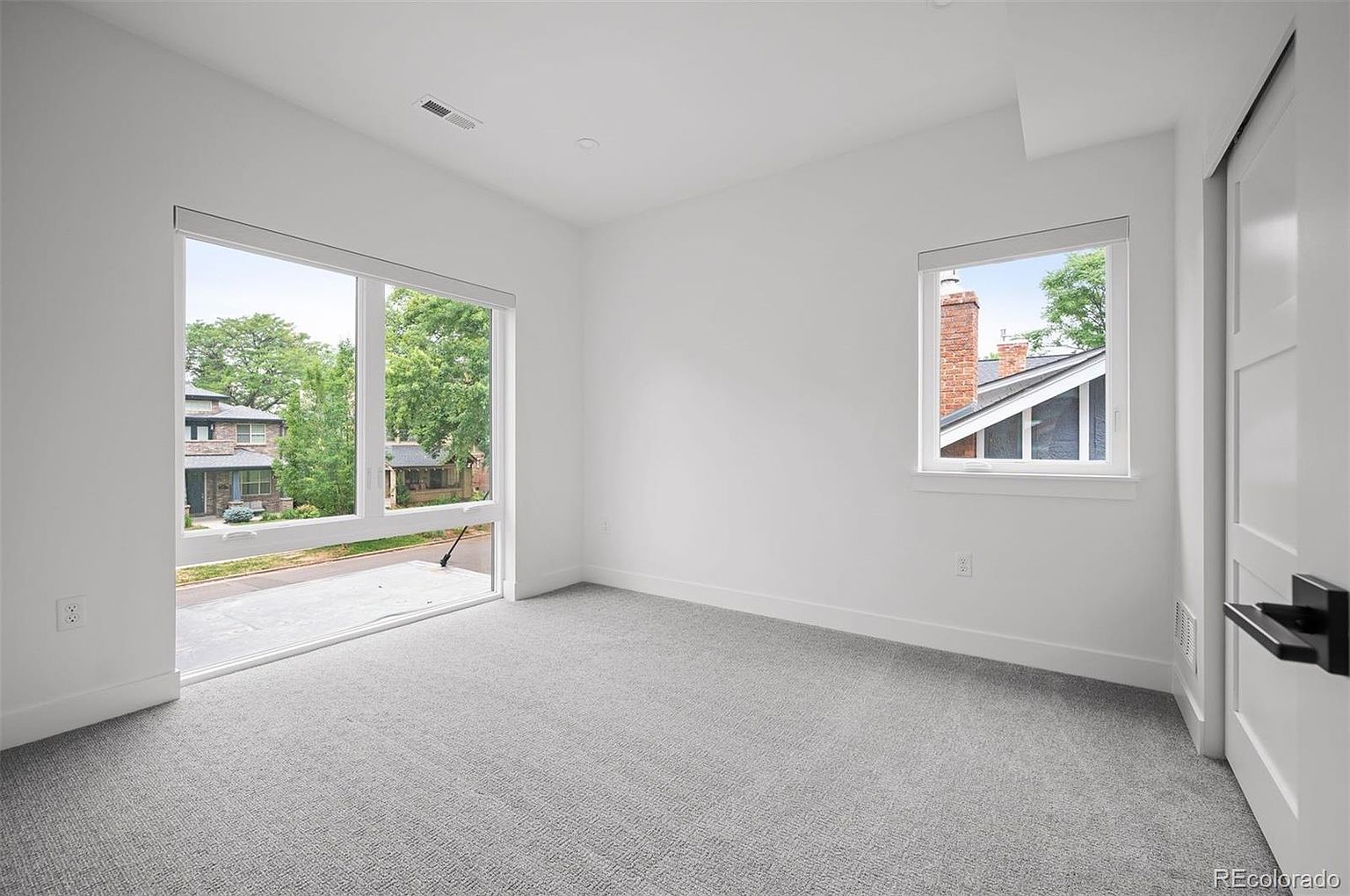 This is a bright, modern bedroom with neutral gray carpet and white walls. A large window offers a view of the neighborhood, while a smaller window provides additional natural light. A sliding door is visible on the right, suggesting a closet or bathroom.