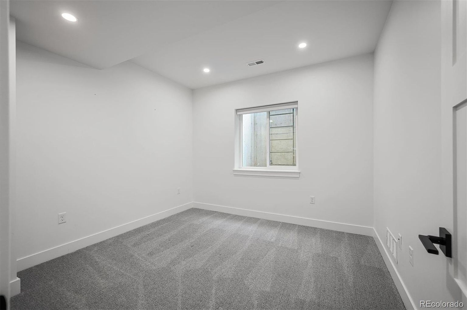 This is an interior shot of a bedroom, likely a guest or secondary bedroom, featuring neutral gray carpet and white walls. A small window provides natural light, and recessed lighting illuminates the room. The overall impression is clean, simple, and ready for personalization.