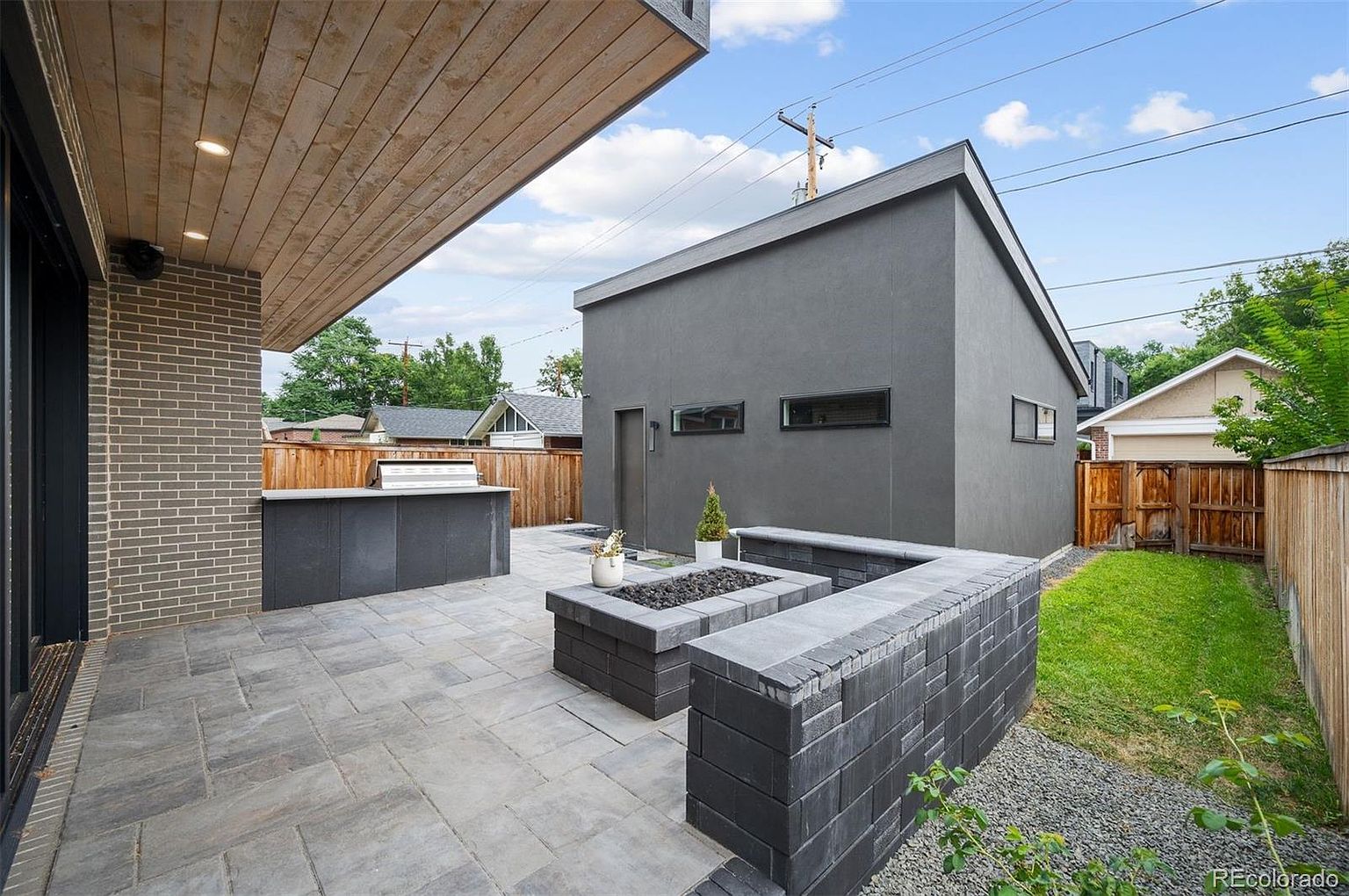 This image showcases a modern patio area with a built-in fire pit and outdoor kitchen. The patio is paved with gray stone tiles, and a gray brick wall surrounds the fire pit. A sleek, gray building stands in the background, complemented by a well-maintained lawn and wooden fence, creating a stylish and inviting outdoor living space.