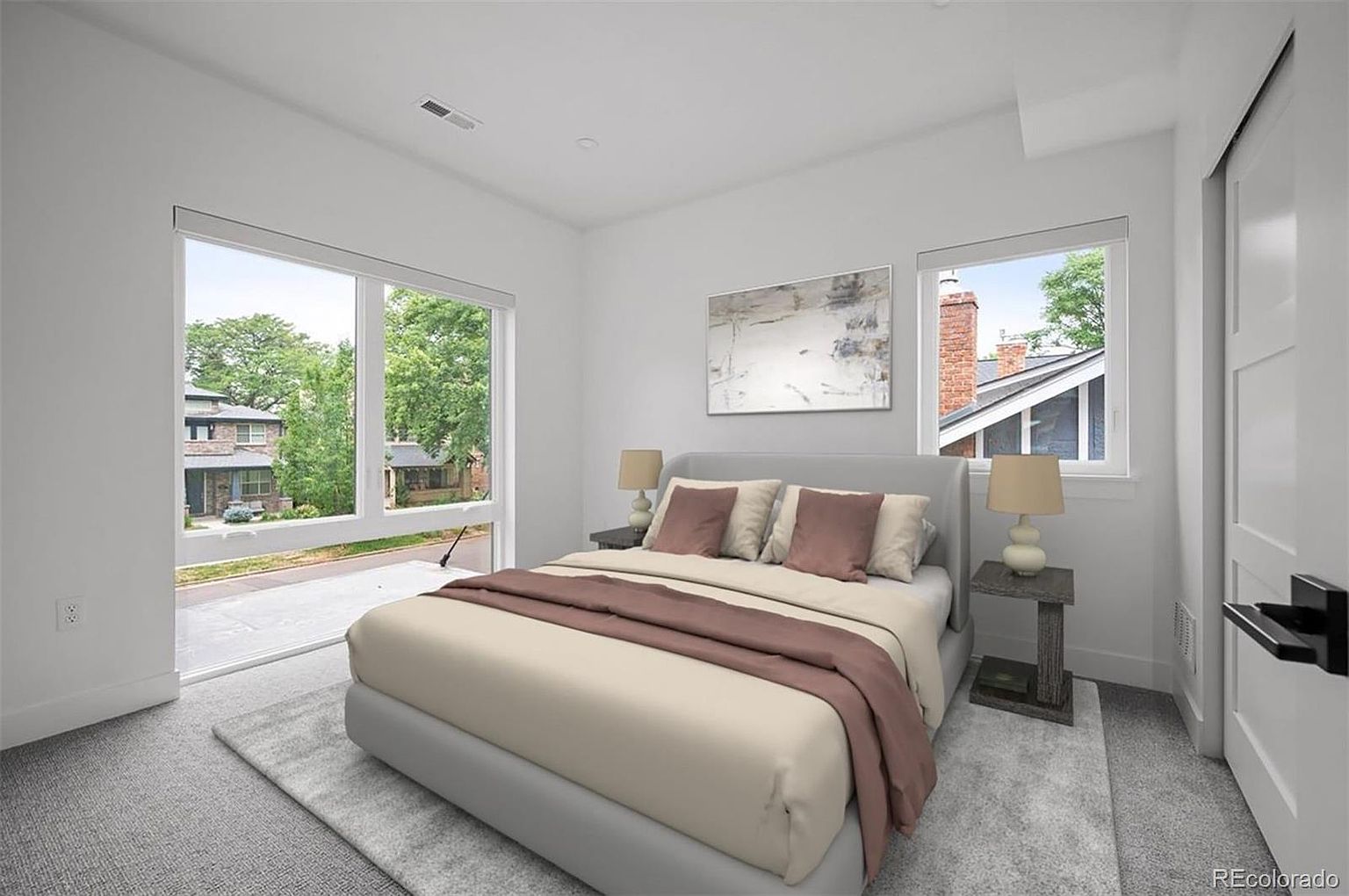 This is a bright and airy primary bedroom featuring a large window with a view of the neighborhood. The room is decorated in neutral tones with a gray bed frame, beige bedding, and brown accents. Two bedside tables with lamps flank the bed, and a piece of artwork hangs above the headboard, creating a serene and inviting atmosphere.