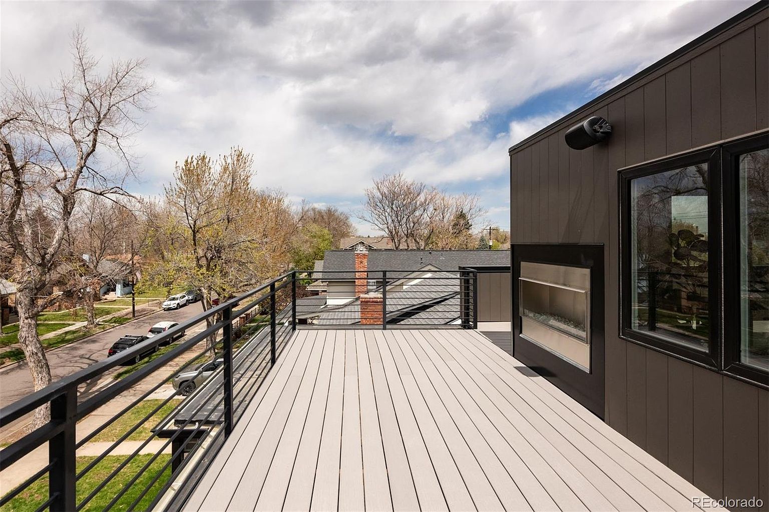 This image showcases a modern rooftop deck with sleek, horizontal railings and light-colored composite decking. A contemporary outdoor fireplace is built into the side of the house, and a large window provides views from inside. The deck overlooks a residential street with mature trees, creating a blend of urban and natural scenery.