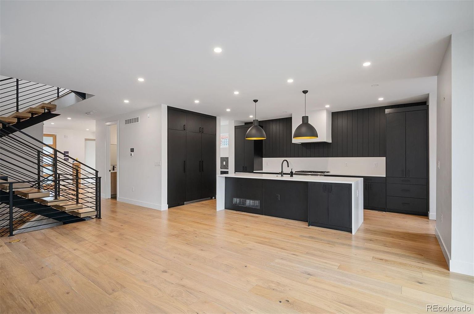 This is a modern, open-concept kitchen and living area featuring dark cabinetry, a large island with a white countertop, and hardwood floors. The kitchen is well-lit with recessed lighting and pendant lights over the island. A staircase with metal railings is visible on the left, adding to the contemporary design of the space.