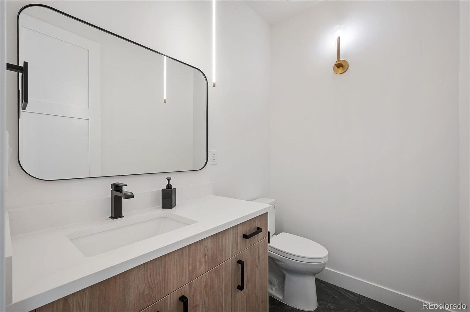 This is a modern guest bathroom featuring a sleek vanity with a white countertop and wood-grain cabinets. A black faucet and soap dispenser complement the black-framed mirror. The walls are painted white, and a stylish gold sconce provides accent lighting, creating a clean and contemporary aesthetic.