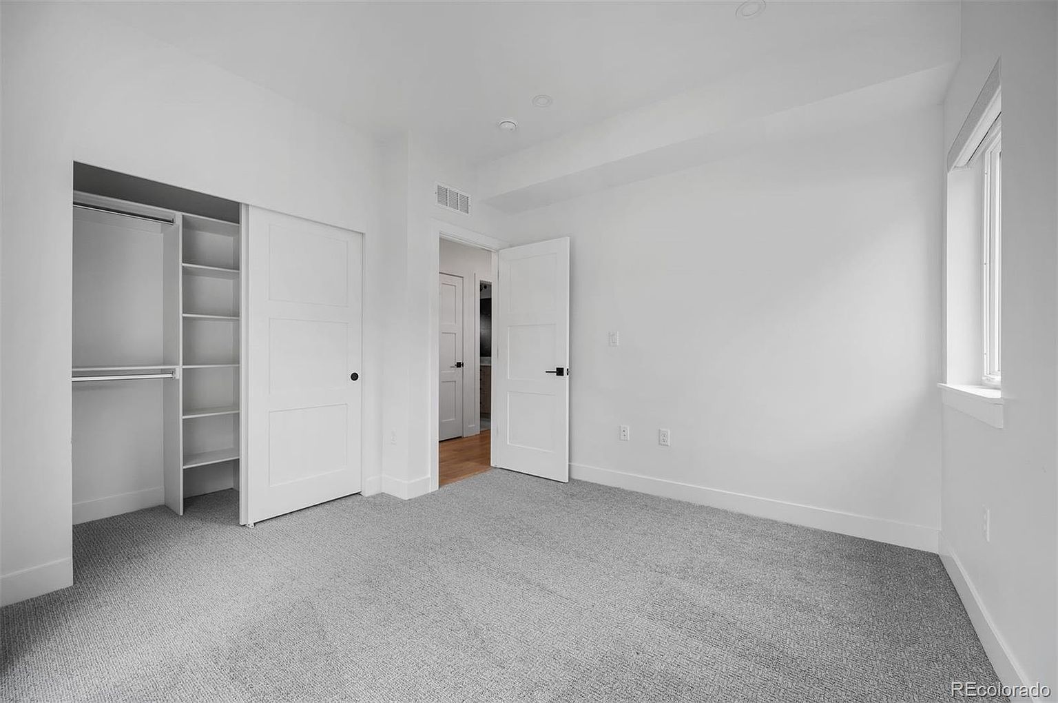 This is a bright and clean bedroom featuring neutral gray carpet and white walls. The room includes a closet with sliding doors and built-in shelving, as well as a doorway leading to another room. A window provides natural light, enhancing the airy feel of the space.