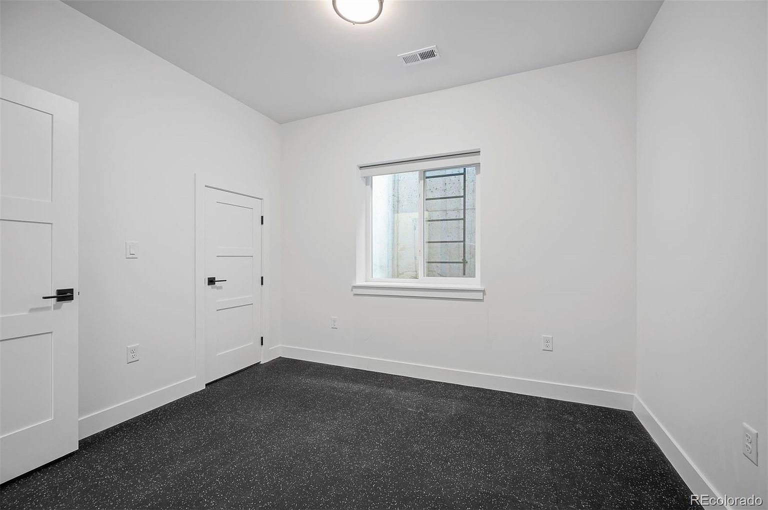 This is a simple, modern bedroom with white walls and a dark gray speckled floor. The room features a window with a view of a concrete wall and ladder, and two white doors with black hardware. The space is clean and minimalist, suitable for a guest room or secondary bedroom.